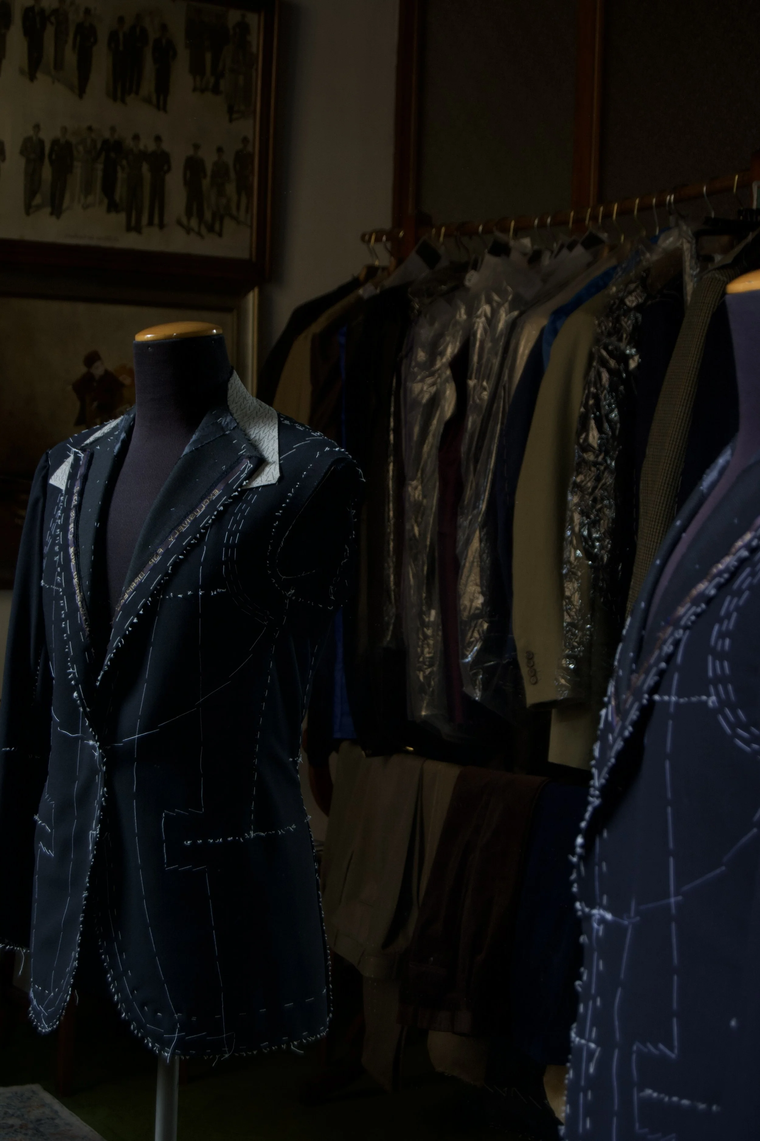 A mannequin dressed in a black suit with white stitching details, positioned in a room with clothing racks and framed artwork on the wall.