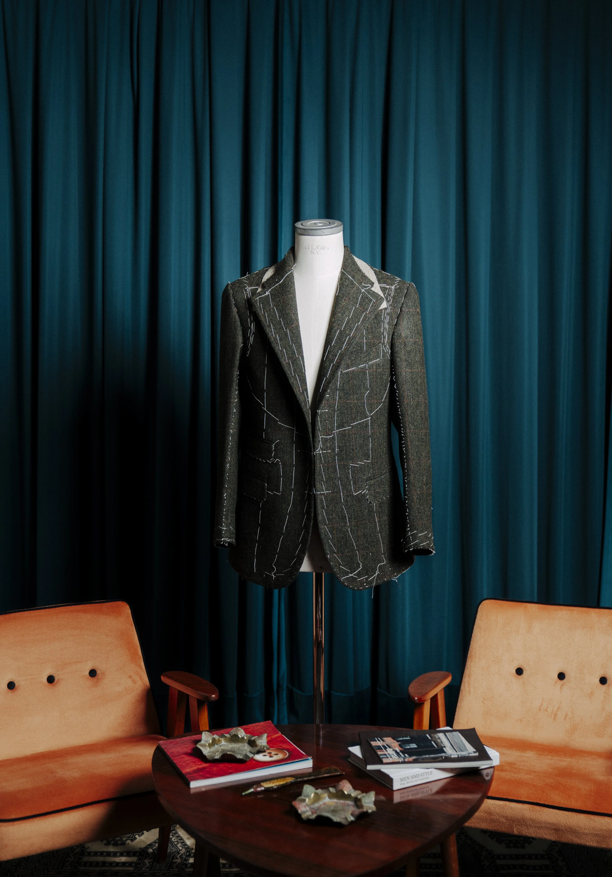 Interior of a room with wooden chairs, a round wooden table with magazines and holiday decorations, and a mannequin dressed in a tailored blazer in front of blue curtains.