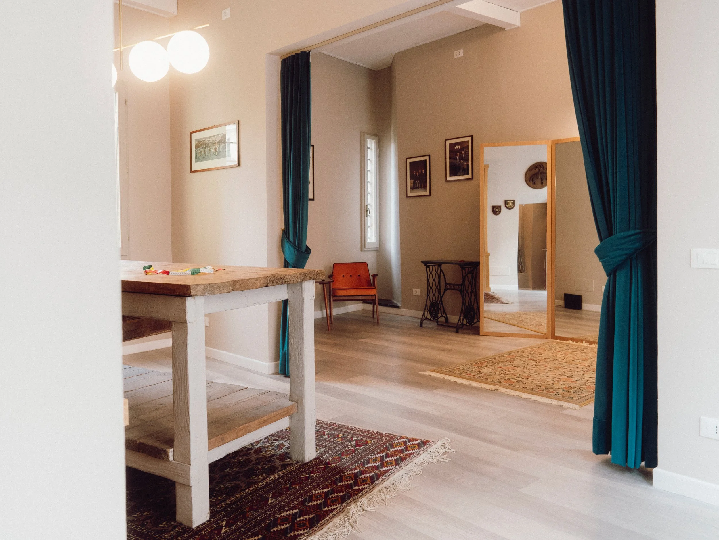 Interior of a living space with a wooden table, framed pictures on the wall, an orange chair, a mirror, blue curtains, area rugs, and natural light coming through windows.