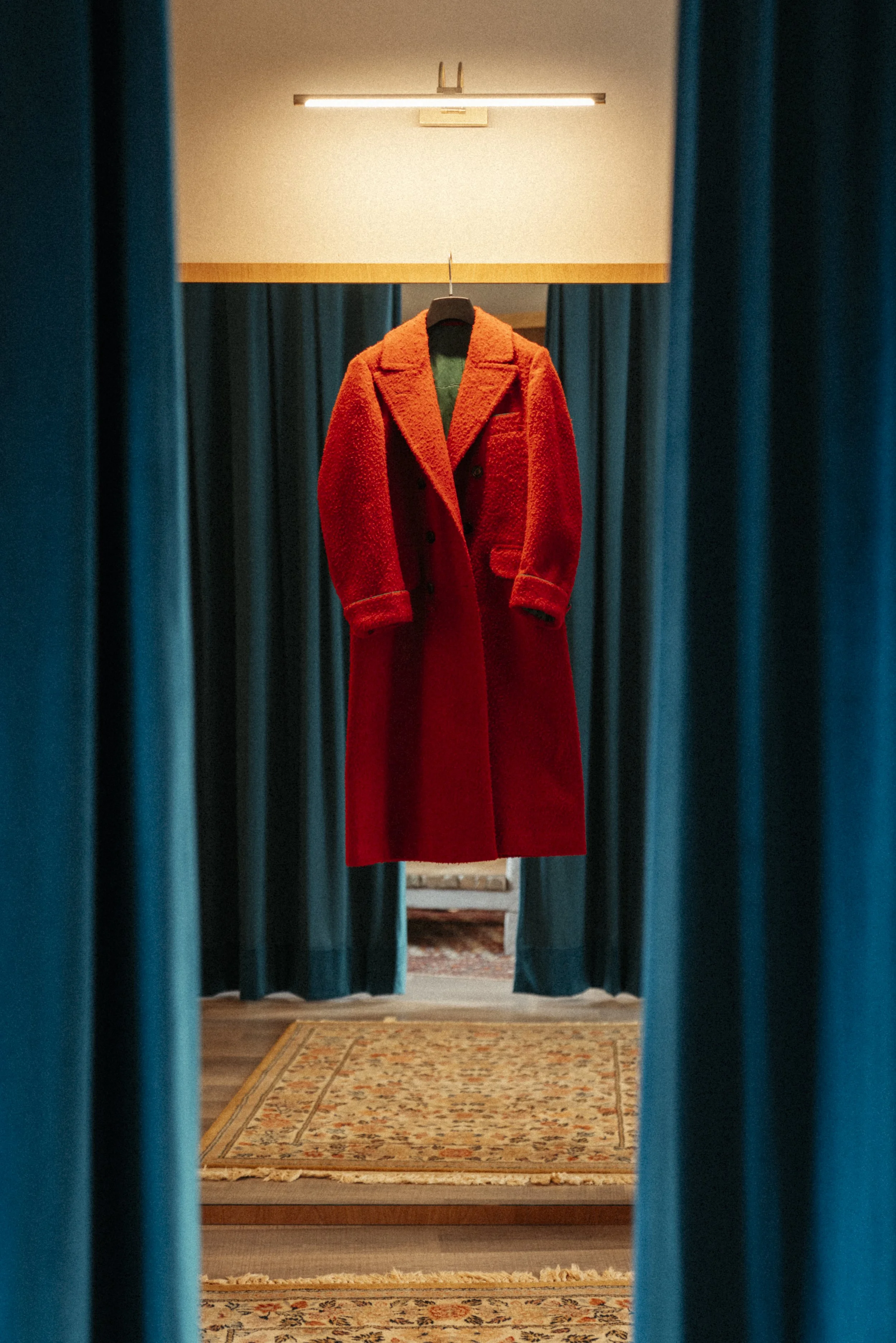 A red coat hanging on a black hanger, viewed through teal curtains with a gold patterned rug and a mirror in the background.