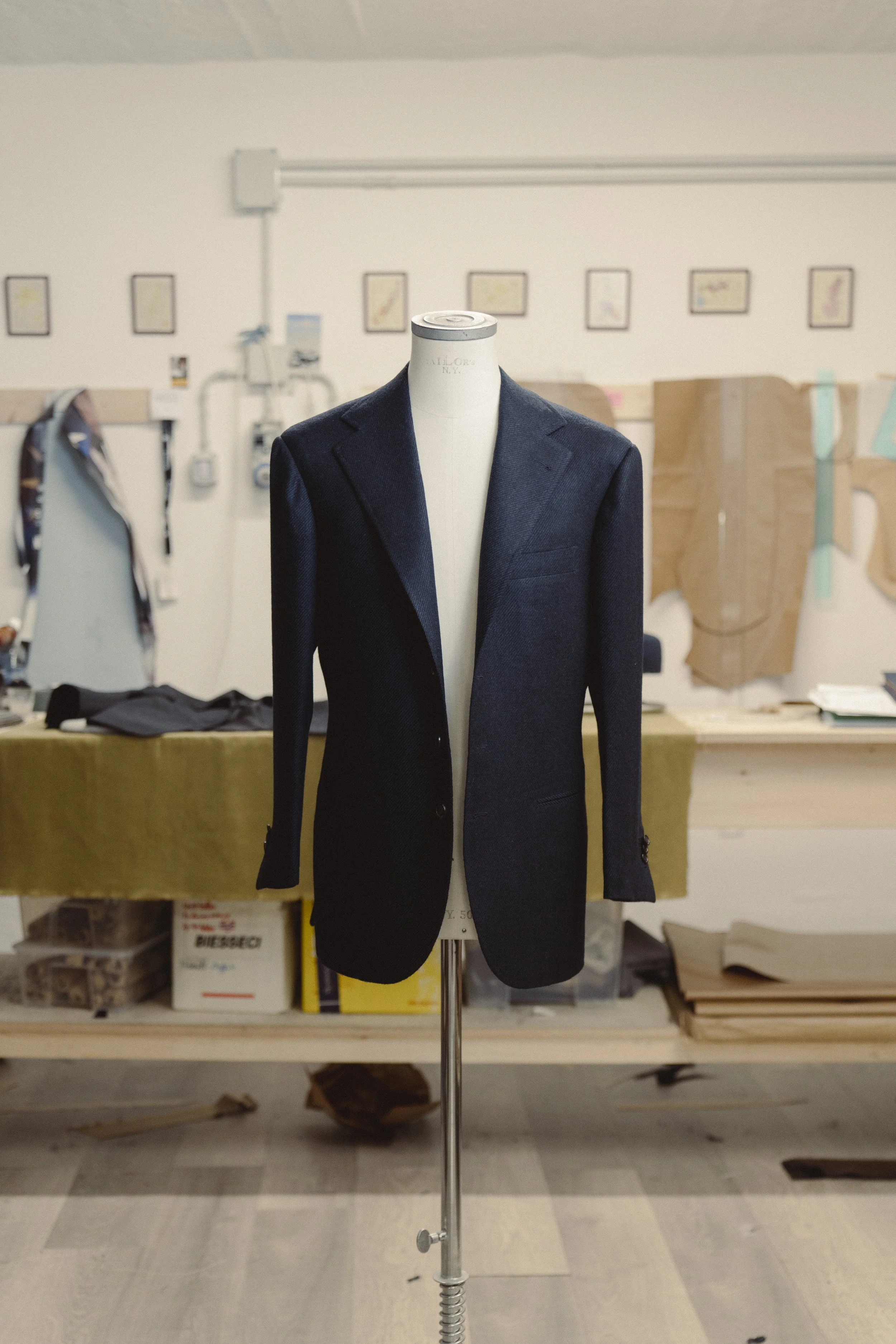A mannequin wearing a dark blue blazer in a workshop or storage room with shelves and boxes in the background.