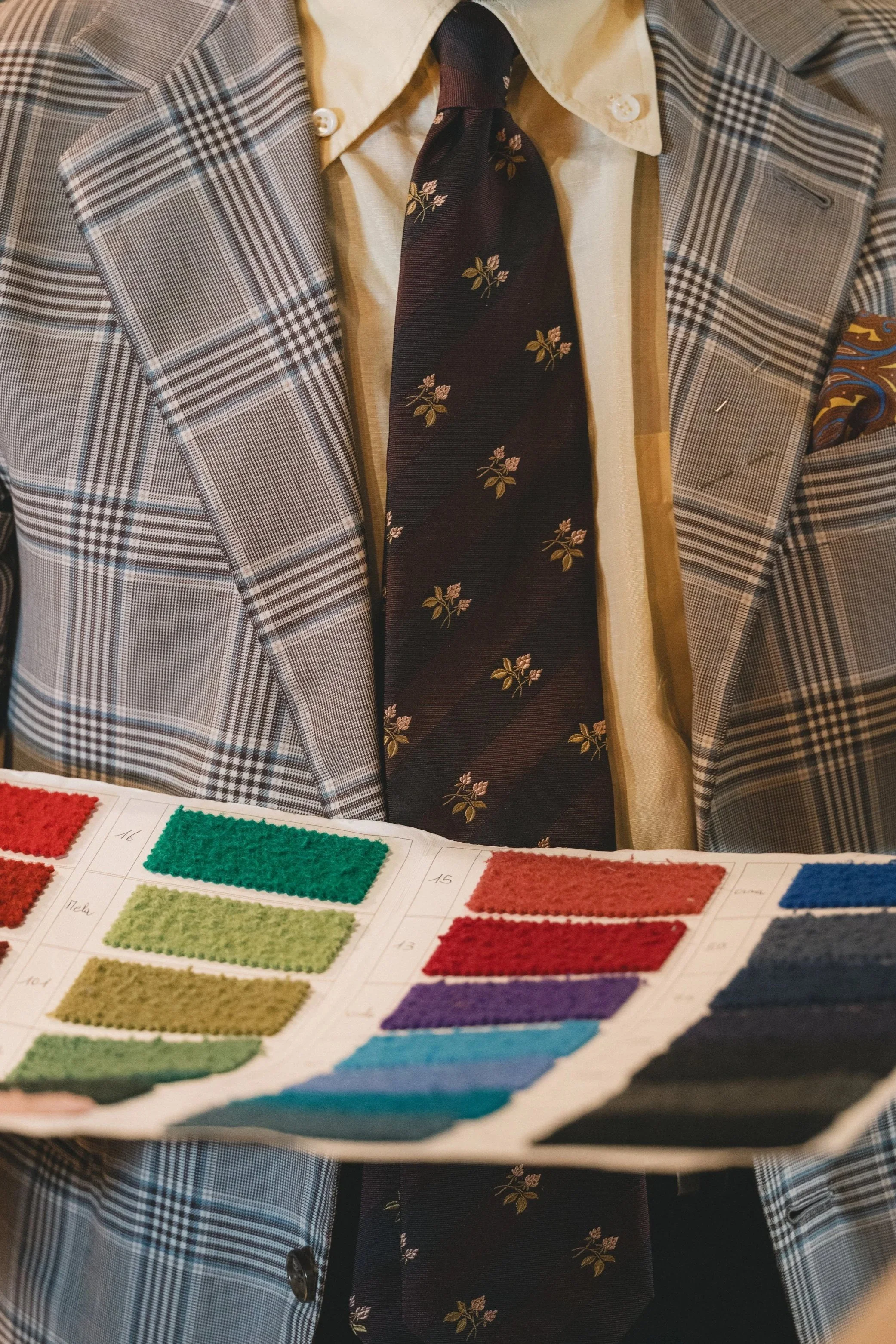 Close-up of a person wearing a checkered blazer, beige shirt, and a dark floral tie, with a color swatch book in the foreground.