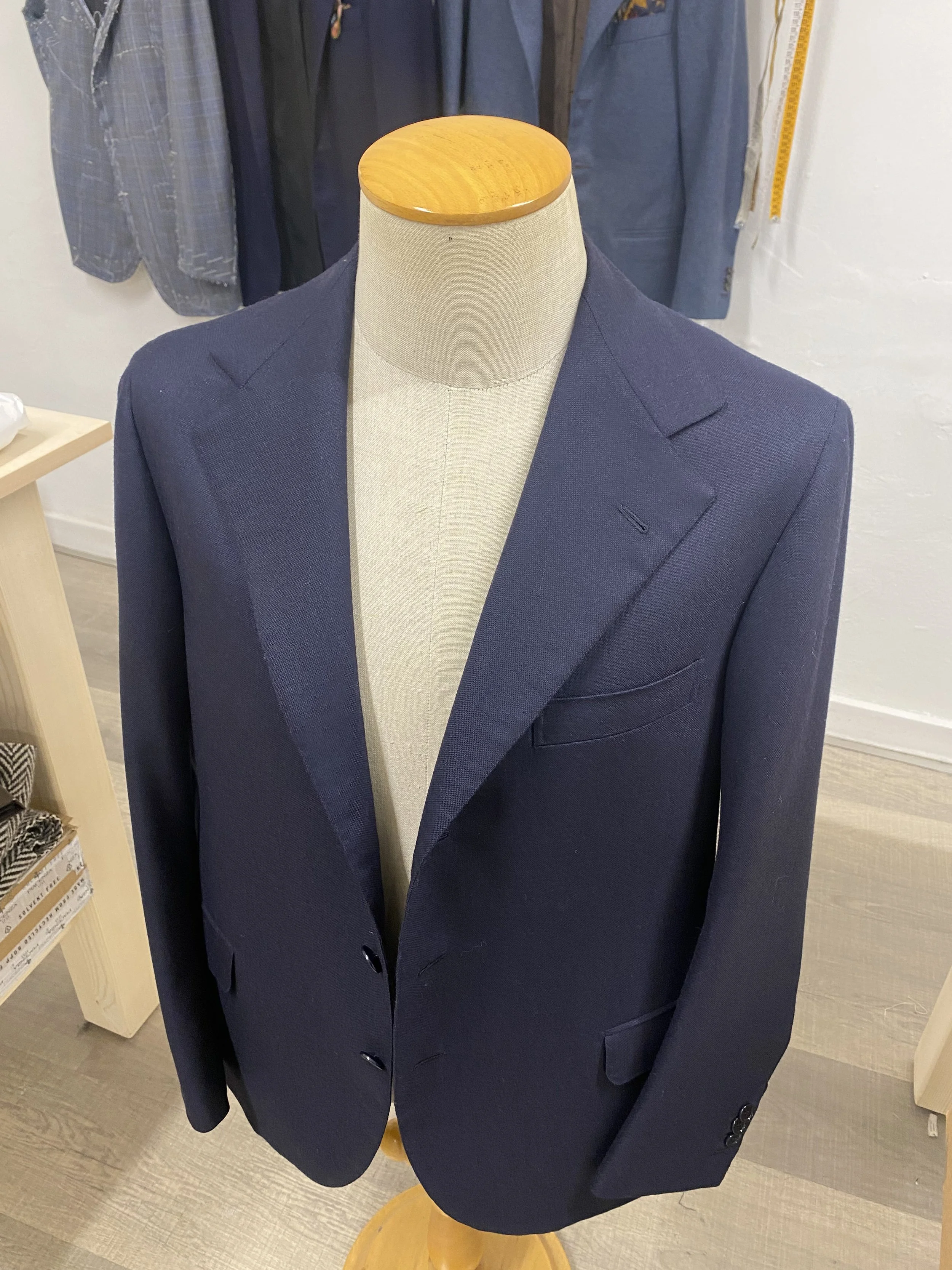 A dark navy blue suit jacket on a beige mannequin torso with a wooden base and no head. The background includes other hanging jackets and a measuring tape on the wall.