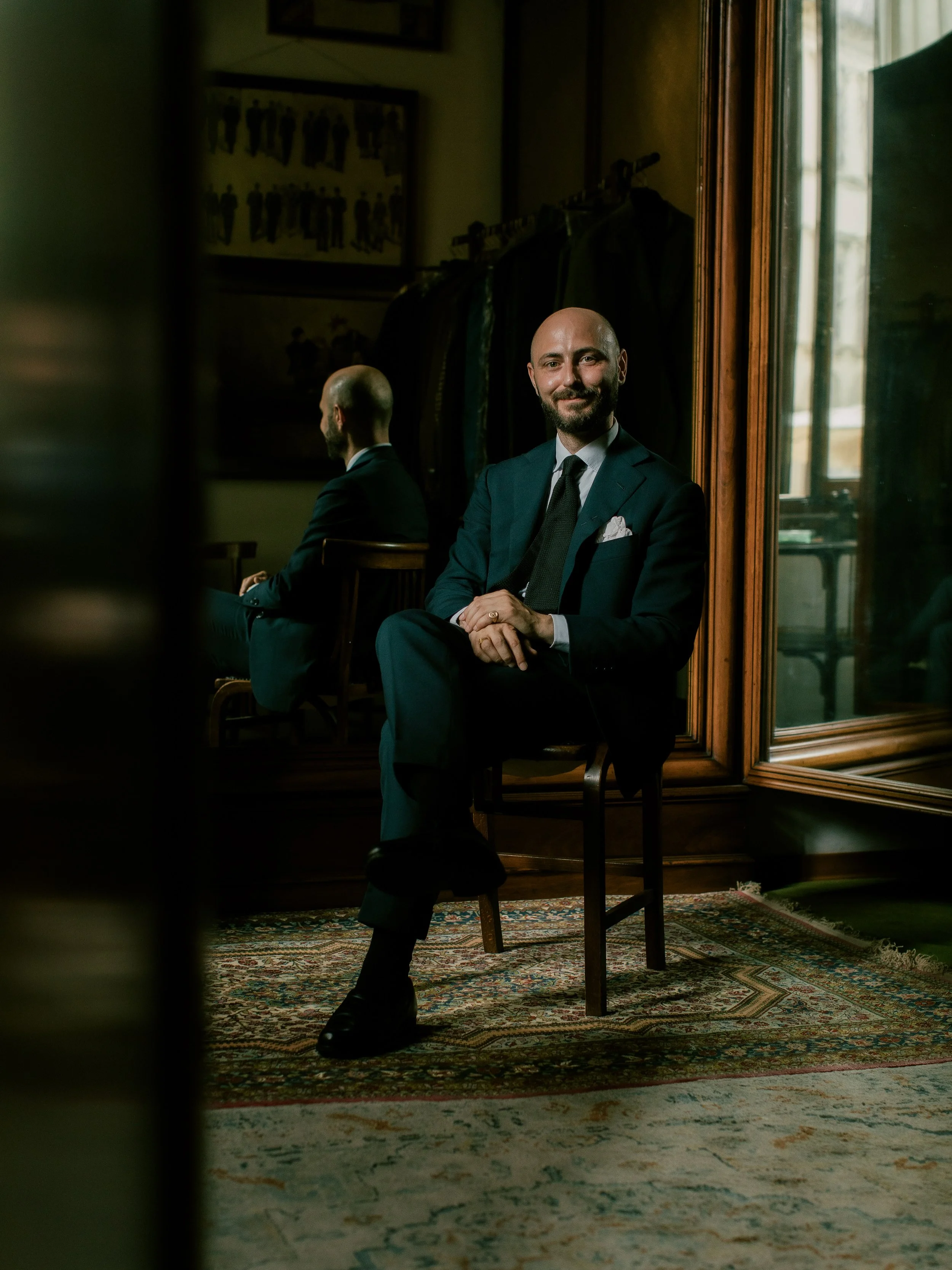 A man in a suit sitting on a chair in a room with a patterned rug, seen through a doorway mirror, with another person reflected in the mirror.