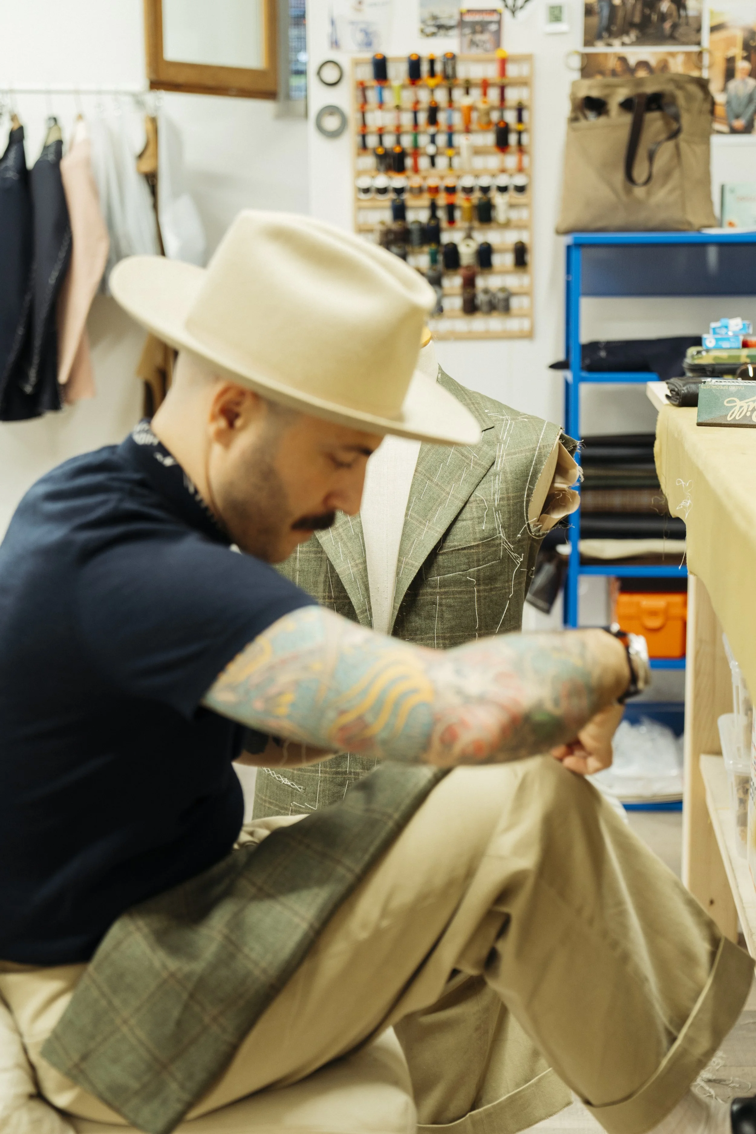 A man with tattoos on his arms working on a tailored suit in a clothing shop, wearing a wide-brimmed hat.