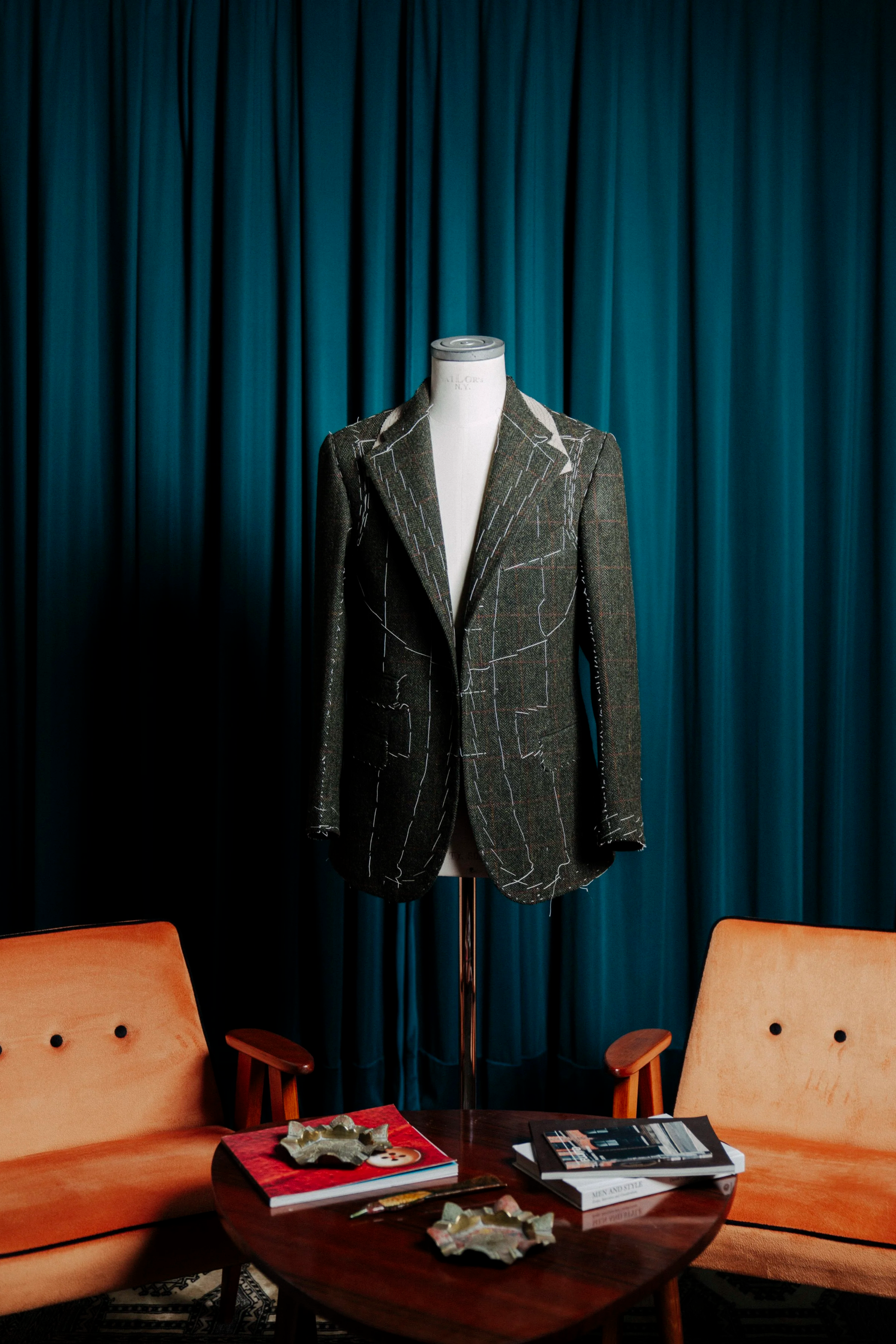 A mannequin wearing a tailored blazer and pants with white stitch outlines, positioned on a stand in front of teal curtains. In front of it is a round wooden table with magazines and scattered decorative items, beside two orange chairs with wooden arms on either side.