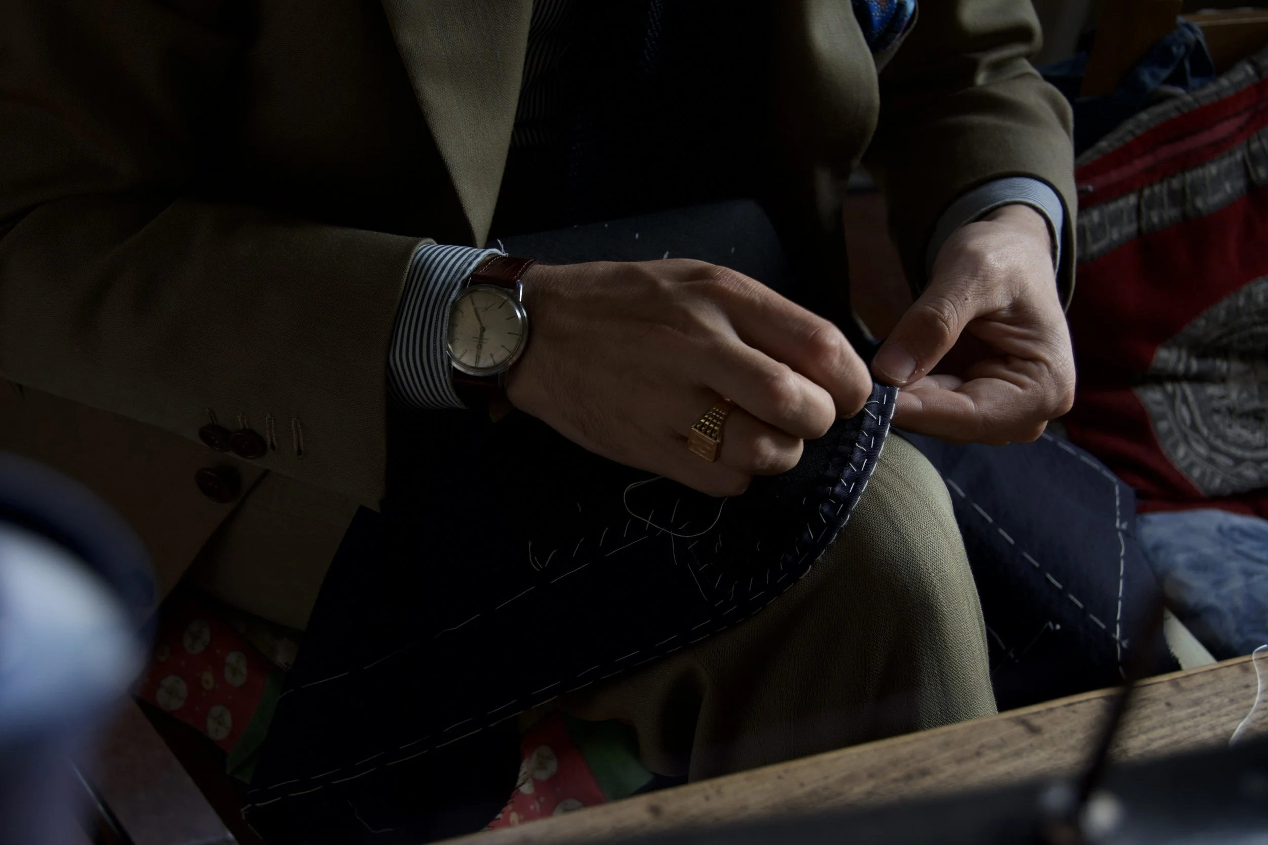 Close-up of a person wearing a tan blazer with a striped shirt cuff, a wristwatch, and a gold ring, holding and working on a small dark blue object with visible stitching, sitting at a wooden table.