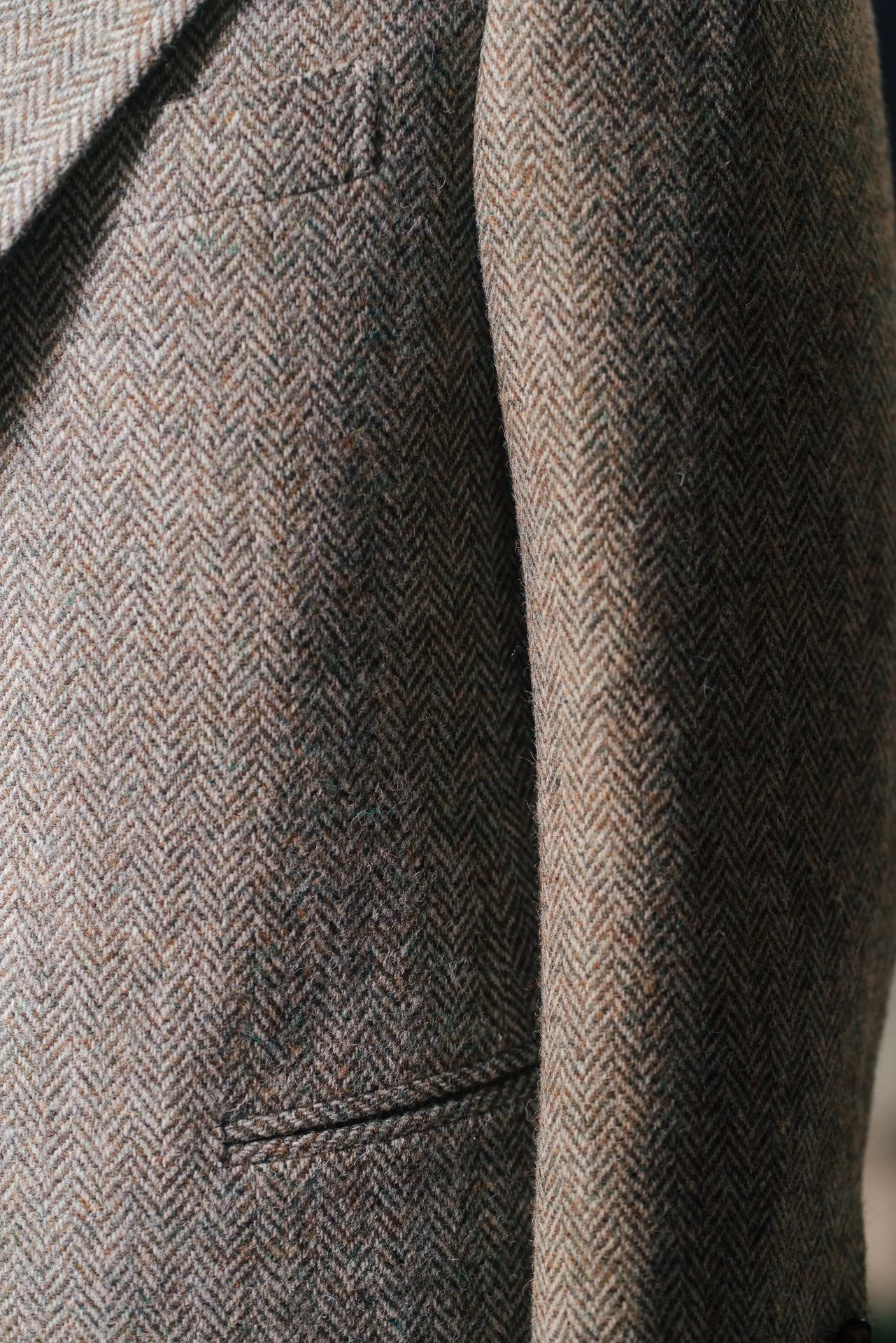 Close-up of a brown tweed blazer with a small pocket on the front, showing the textured herringbone pattern of the fabric.