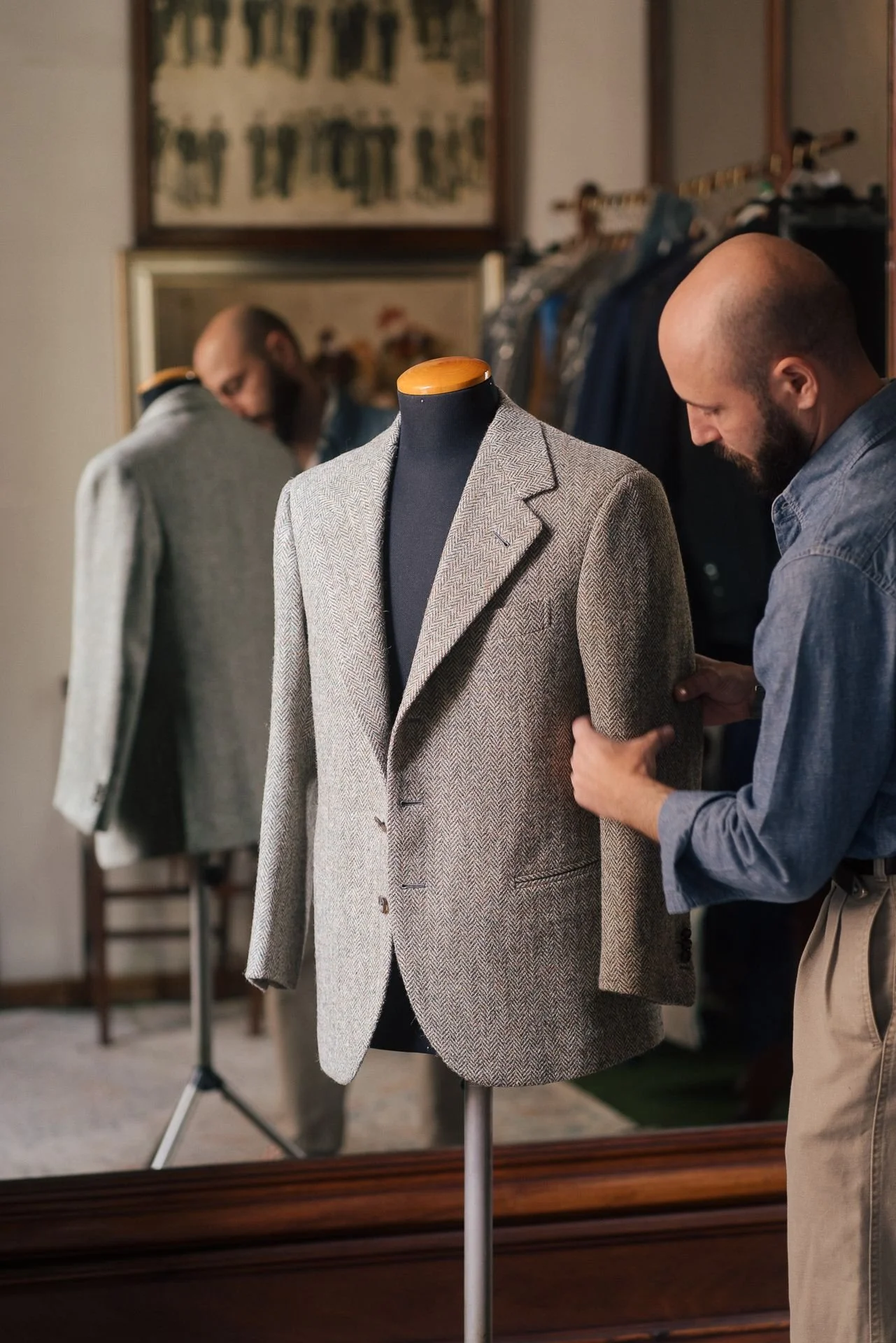 A man trying on a beige checkered blazer in front of a mirror, with clothing rack and artwork on the wall in the background.