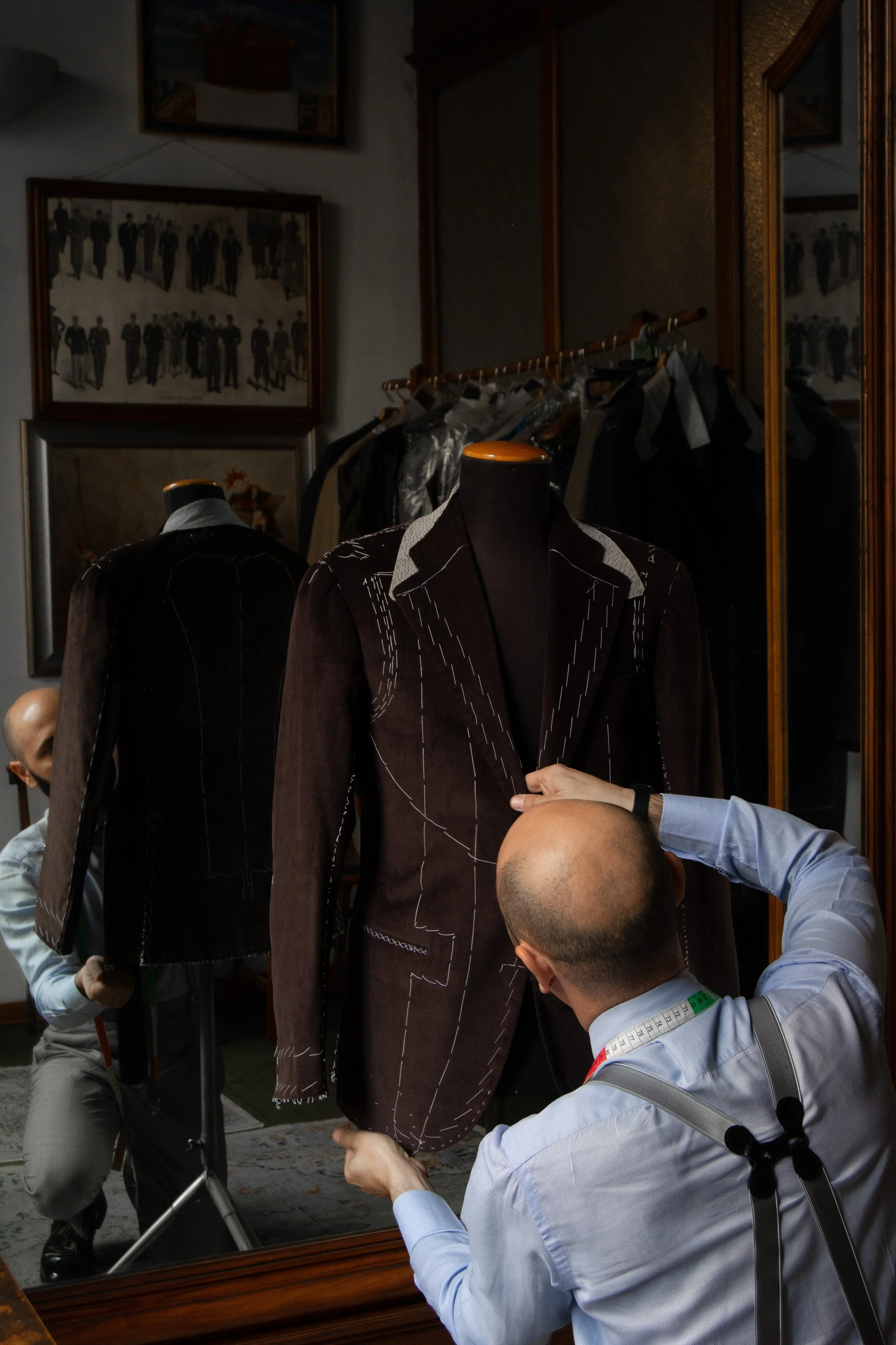 A tailor measures a brown suit jacket on a mannequin while another man adjusts it. In the background, there are framed pictures on the wall, a coat rack with clothing, and a mirror reflecting part of the scene.
