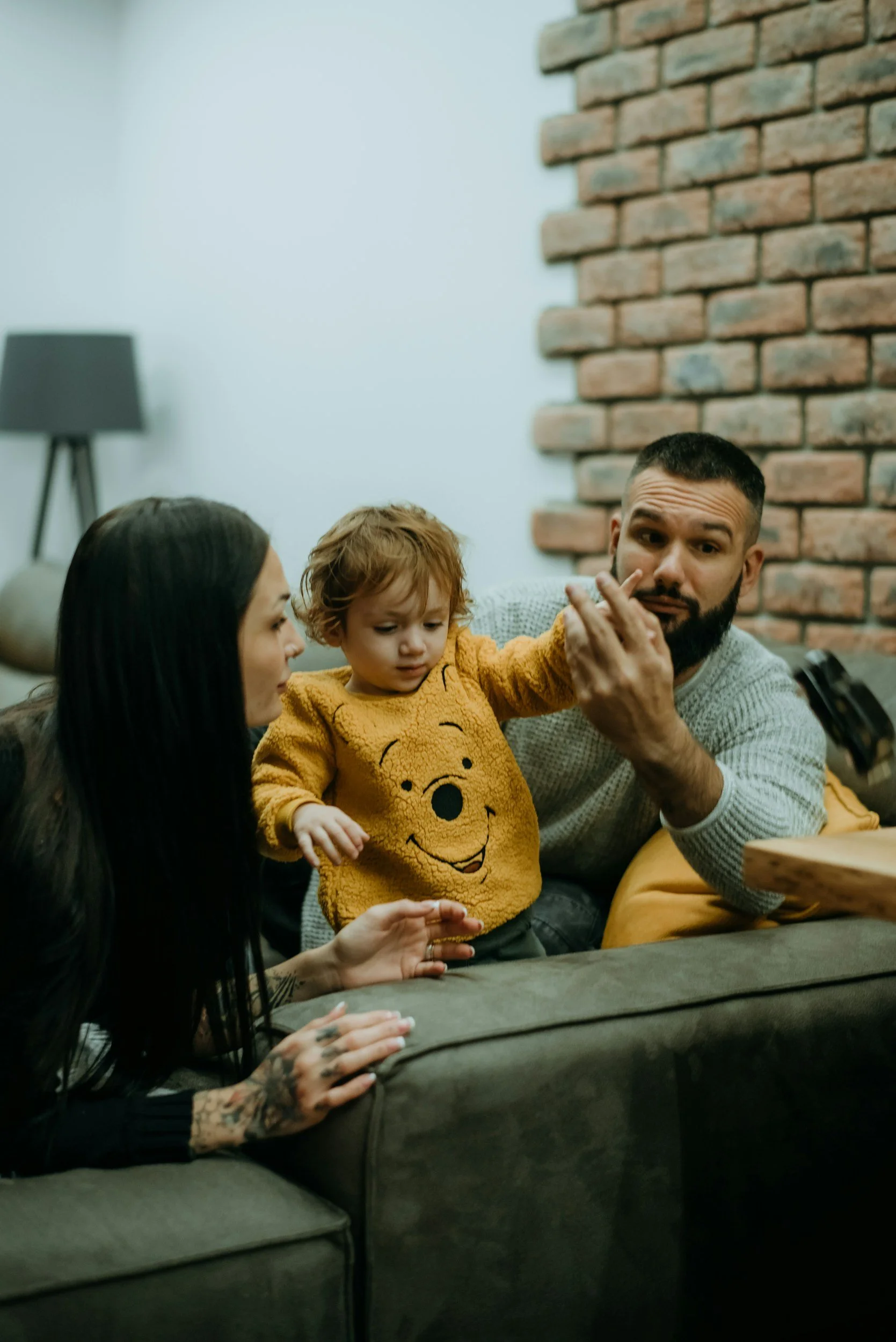 A family gathering with a woman, a man, a young child, and a woman with tattoos, sitting on a couch in a cozy living room with exposed brick wall and a lamp in the background.