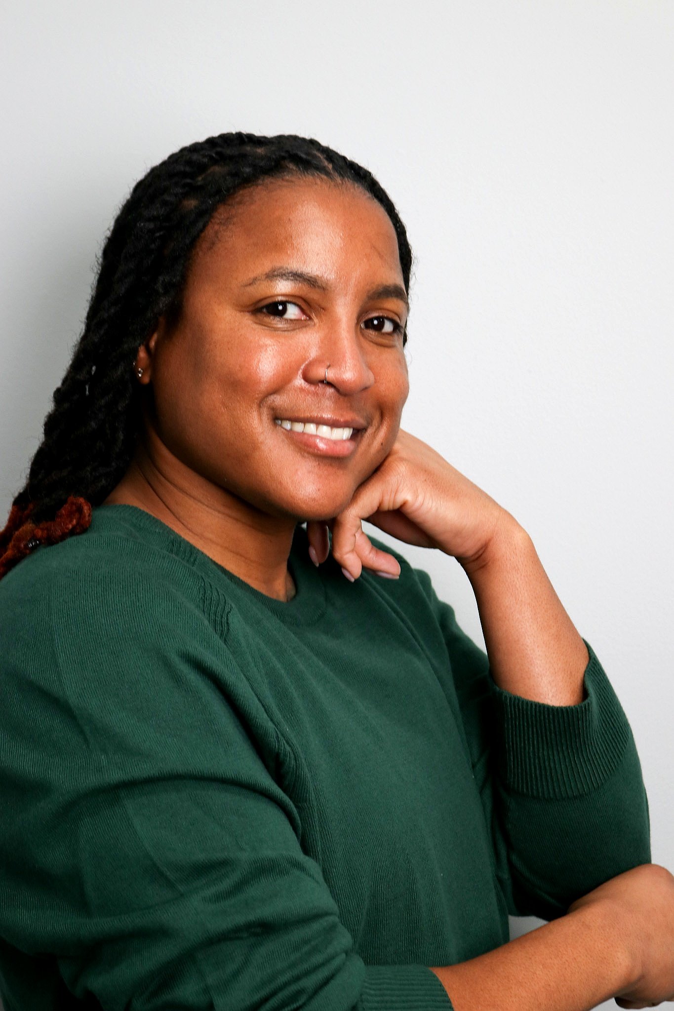 A woman with medium skin, dark braided hair, and a nose ring, smiling and resting her chin on her hand, wearing a green sweater, in front of a plain white background.