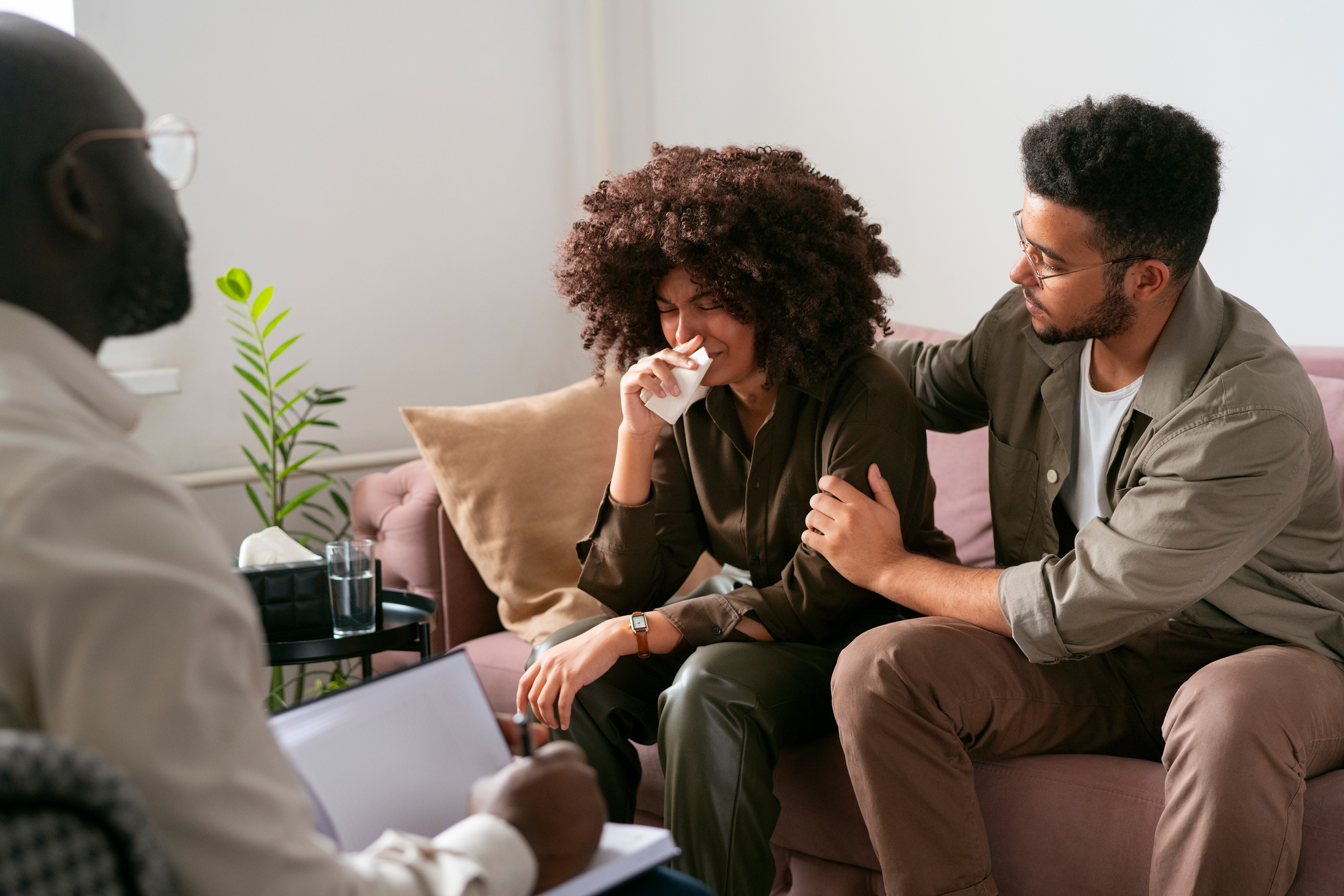 A woman with curly hair appears to be crying while a man comfort her during a therapy or counseling session. Another person, possibly a therapist, is partially visible in the foreground, taking notes.