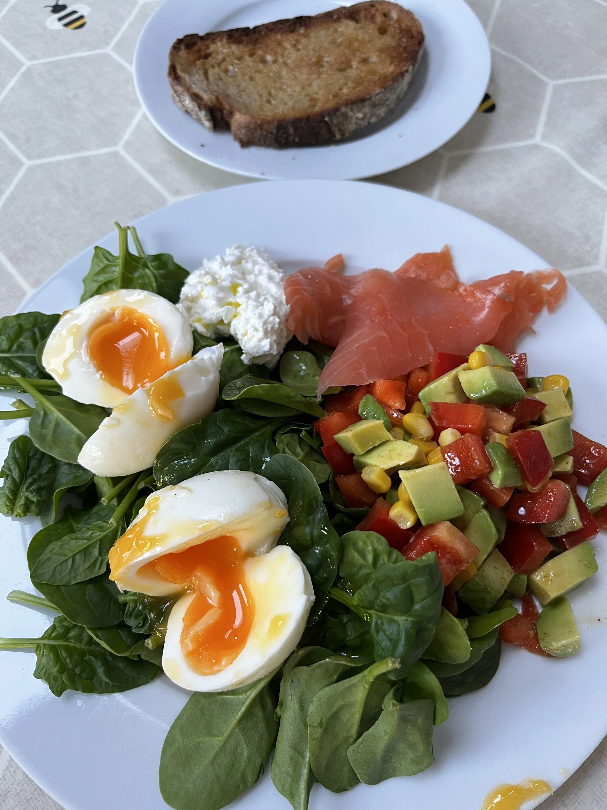 A plate of fresh spinach leaves topped with soft-boiled eggs, shredded cheese, and chopped vegetables including avocado, tomato, and corn. A slice of toasted bread is on a separate plate in the background.