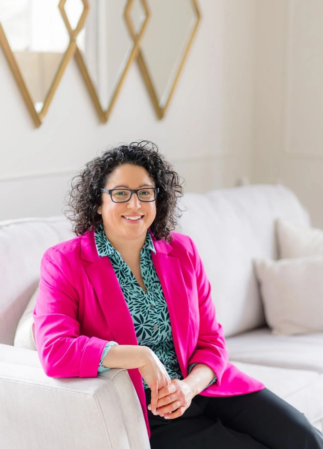 A woman with curly dark hair, glasses, wearing a bright pink blazer and a blue and black patterned blouse, sitting on a white sofa, smiling, in a modern, well-lit room.