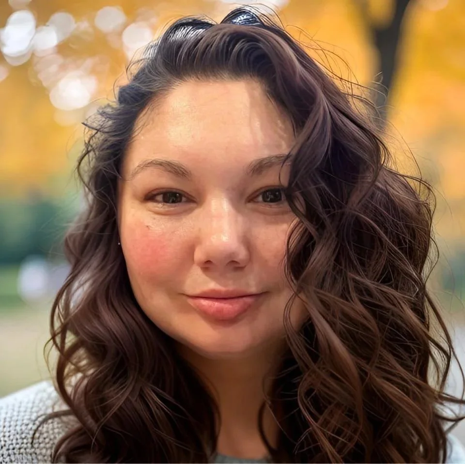 A close-up of a young woman with long, wavy dark hair, smiling softly outdoors during fall with colorful trees in the background.