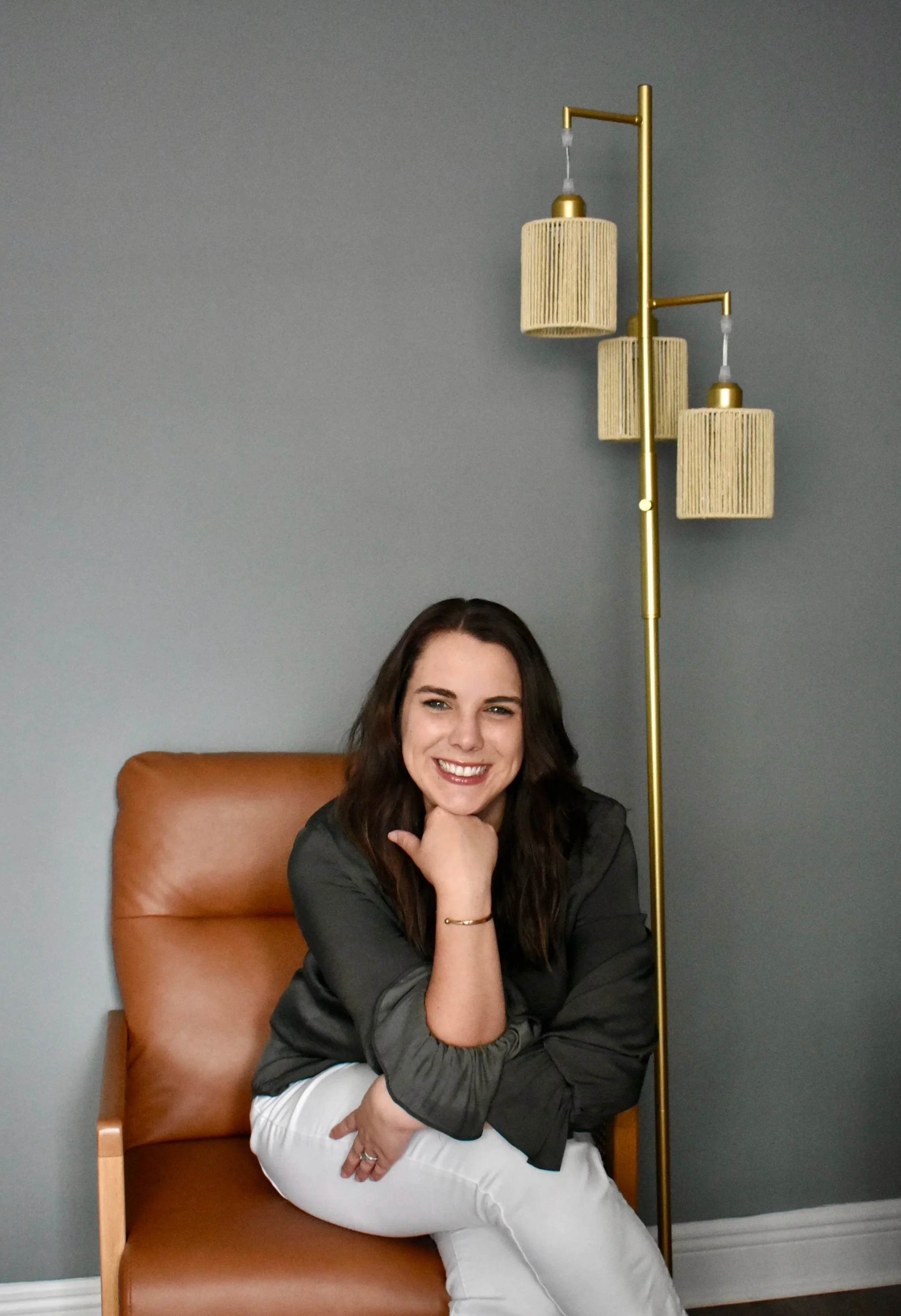 A woman with long dark hair and fair skin, smiling and sitting on a tan leather chair against a gray wall. She is wearing a dark jacket and white pants, with her left hand resting on her chin and her right arm crossed over her lap. A tall, gold-colored floor lamp with three cylindrical, fabric lampshades stands behind her to her right.