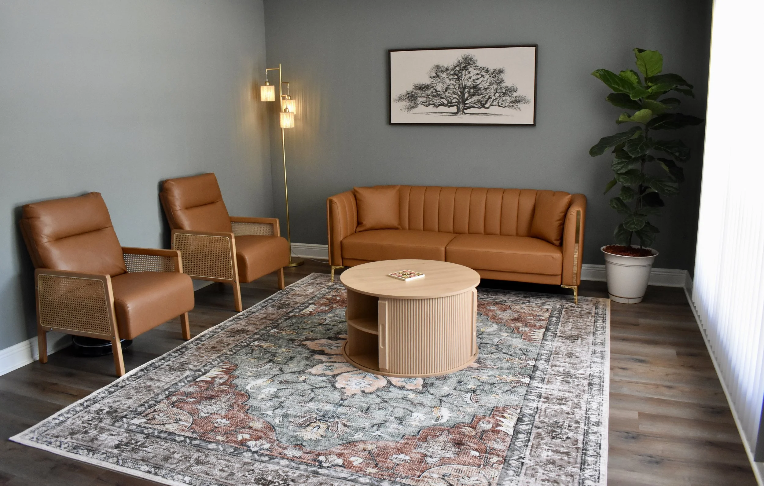 A waiting room with two tan armchairs, a matching sofa, a round wooden coffee table, a patterned area rug, a large potted plant, a framed black-and-white tree print on the wall, a multi-light floor lamp, and a window with vertical blinds.