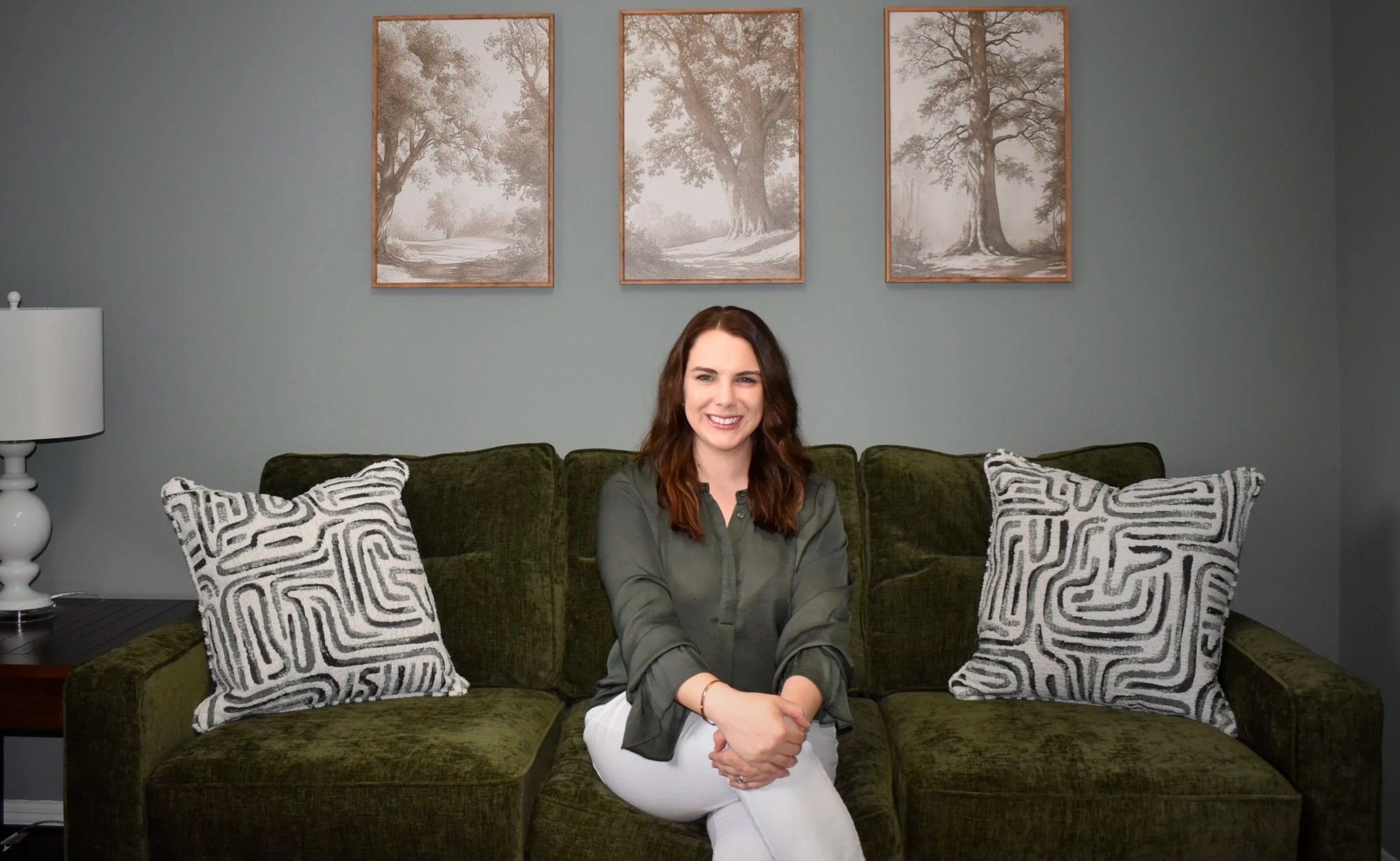 A woman with long brown hair sits on a green velvet sofa, smiling, with three patterned pillows. Behind her, three framed monochrome tree prints hang on a light gray wall. To her left, a white table lamp sits on a small side table.