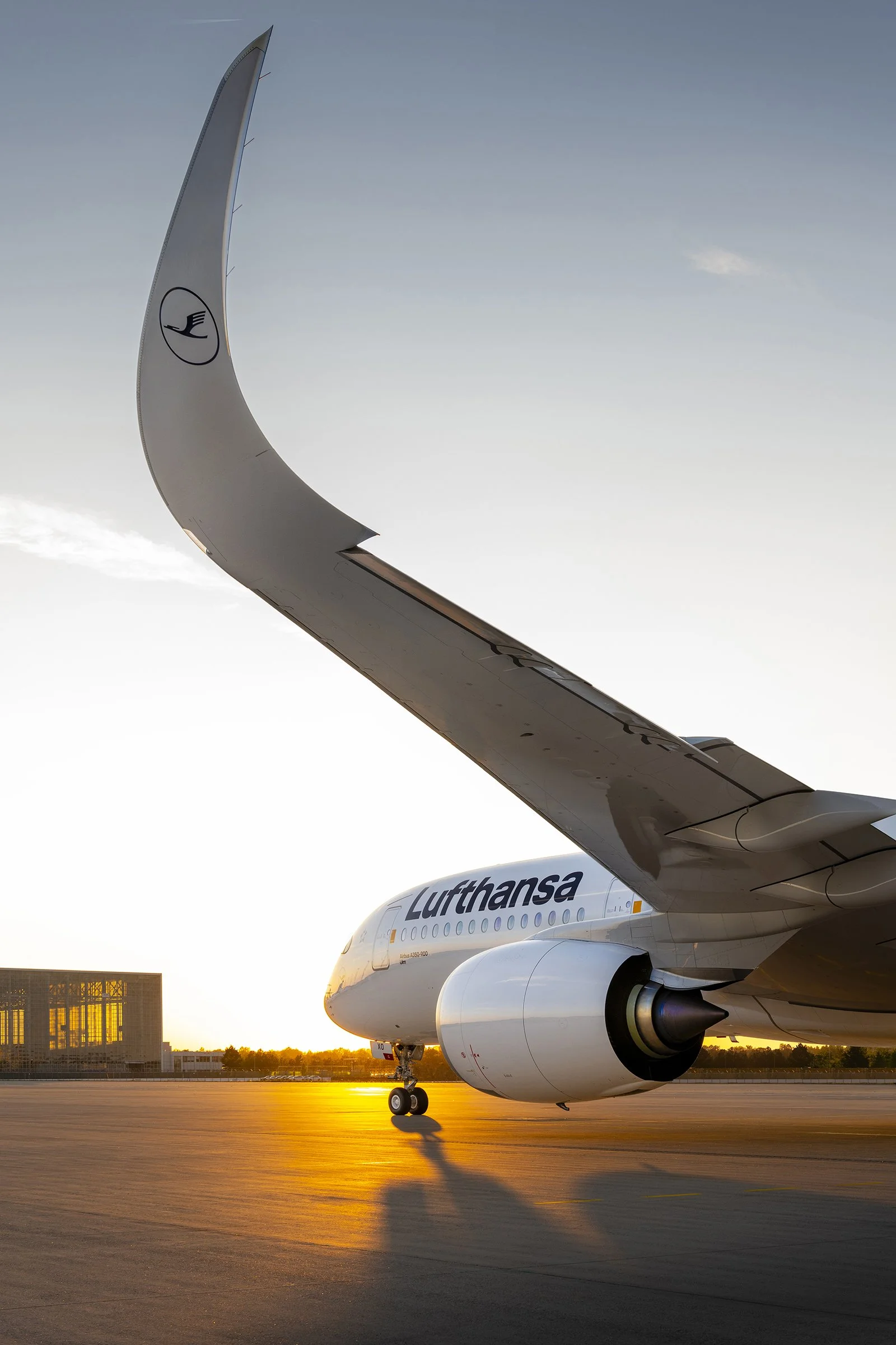 Photo of a Lufthansa airplane on the tarmac at sunset, with the airplane's wing and engine in the foreground and a building and trees in the background.