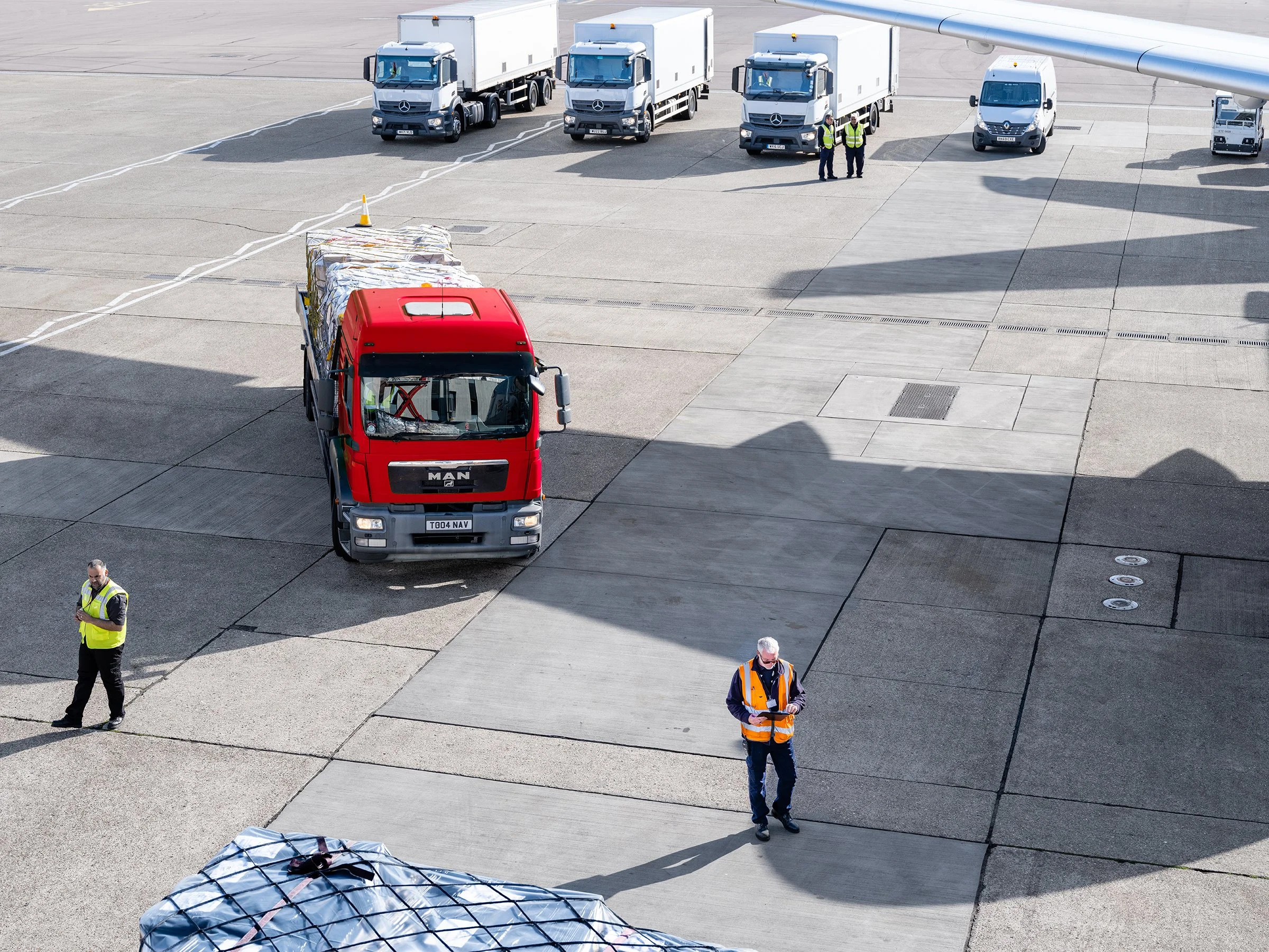 Airport tarmac with luggage carts, trucks, and personnel during daytime.