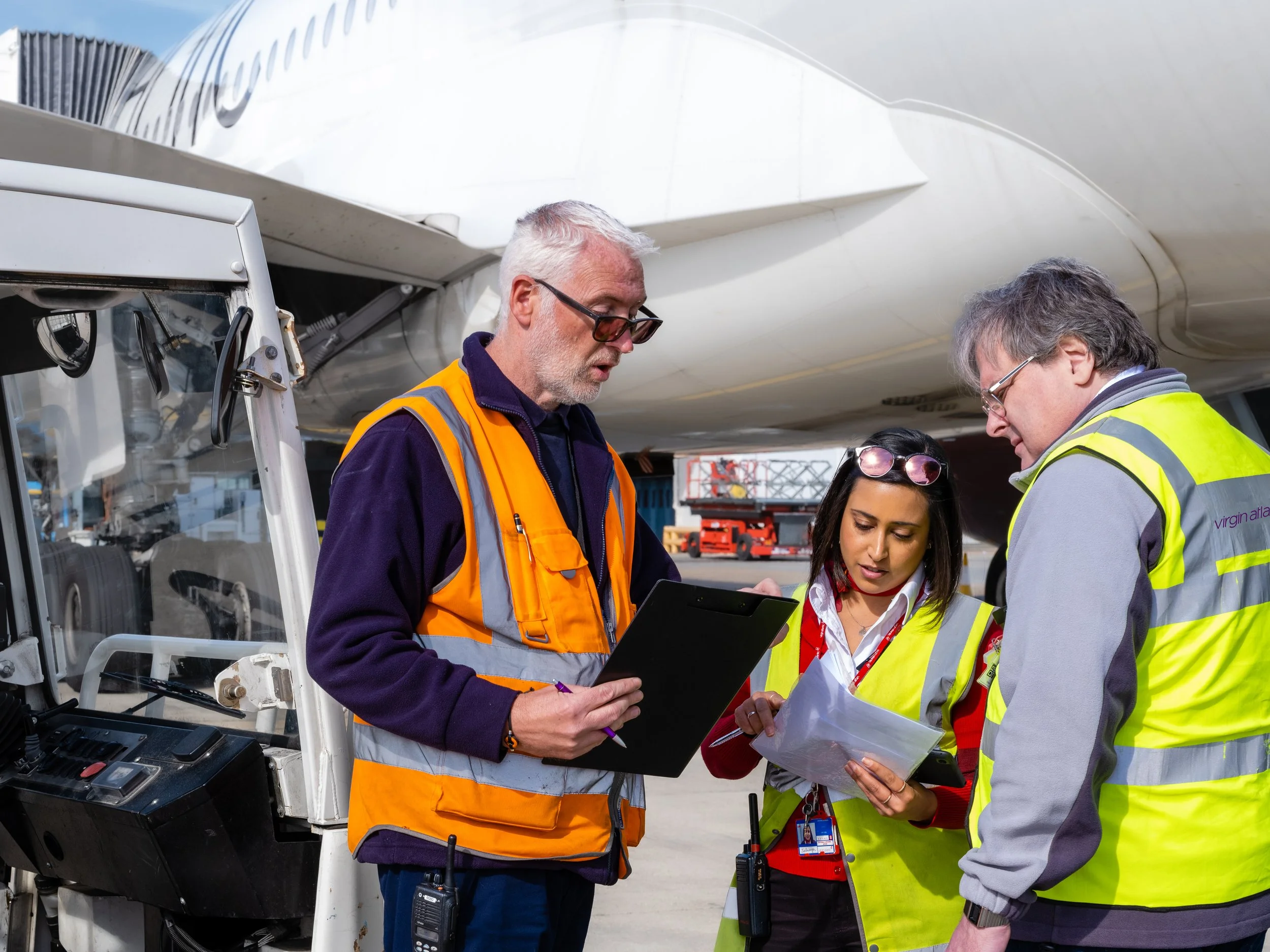 Group of aviation workers in safety vests discussing near an airplane's nose, with equipment and airport background visible.