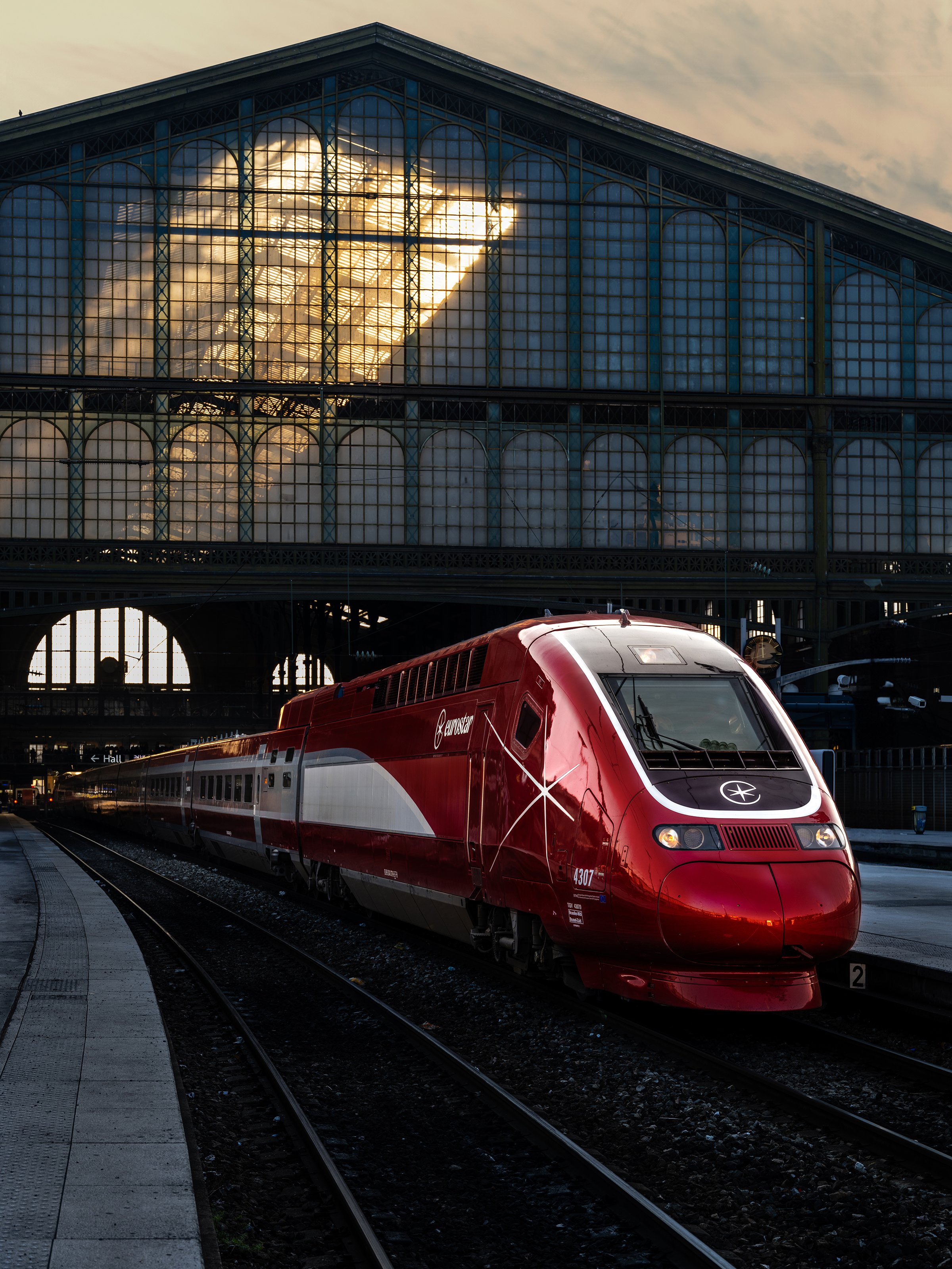 A red high-speed train parked at a train station with a large glass building reflecting the sunset in the background.