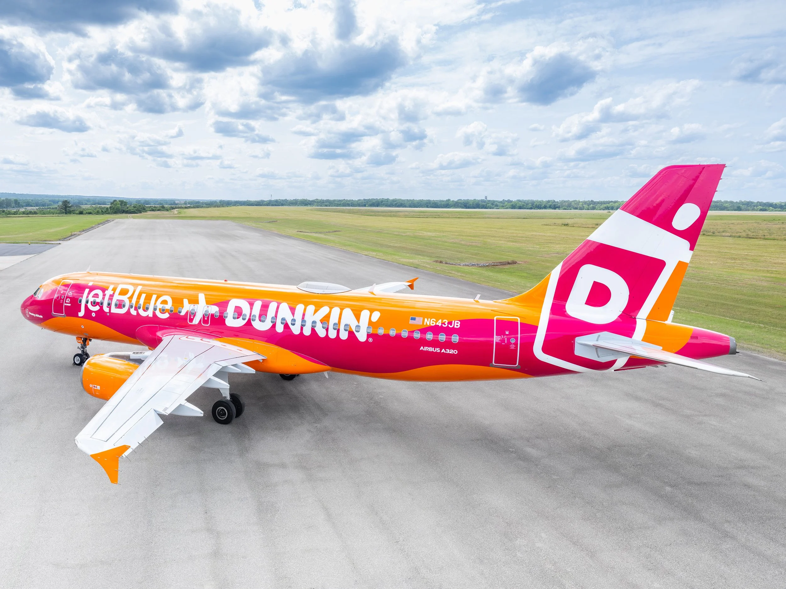 Close-up of a JetBlue airplane with pink, orange, and white livery parked on the runway under a partly cloudy sky.