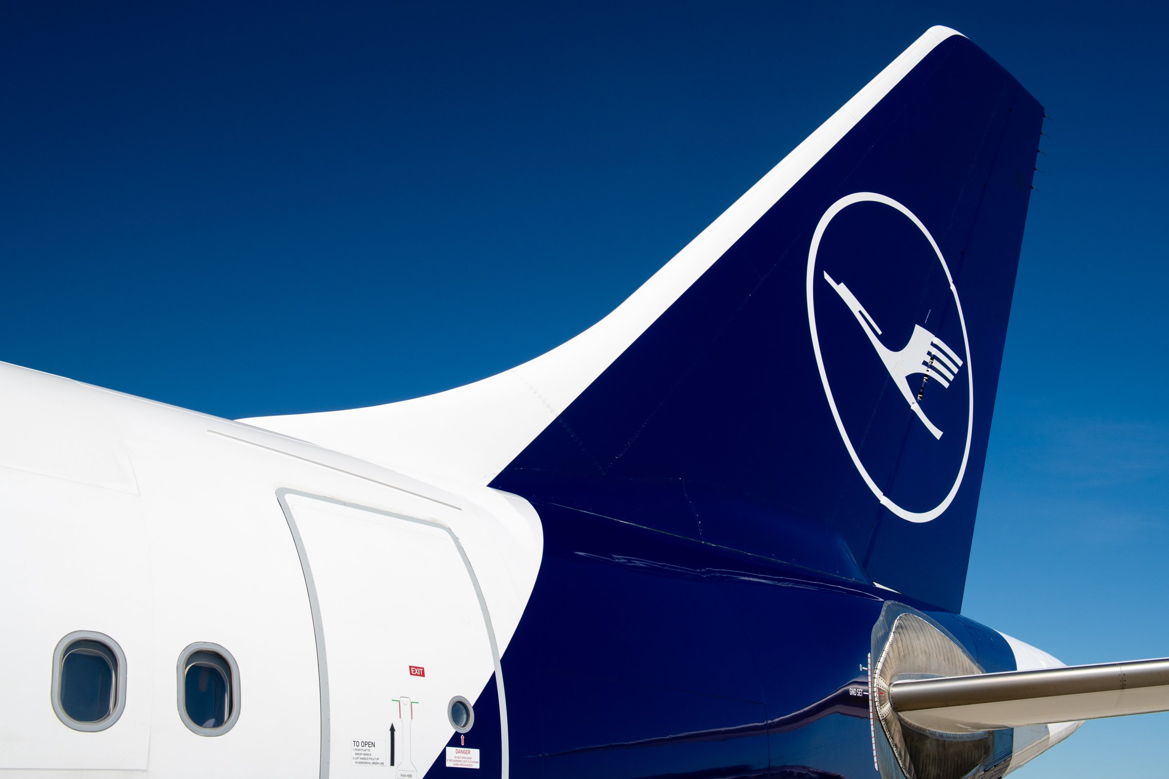 Close-up of the tail section of a blue and white commercial aircraft with a logo of a stylized white circle and line, against a clear blue sky.
