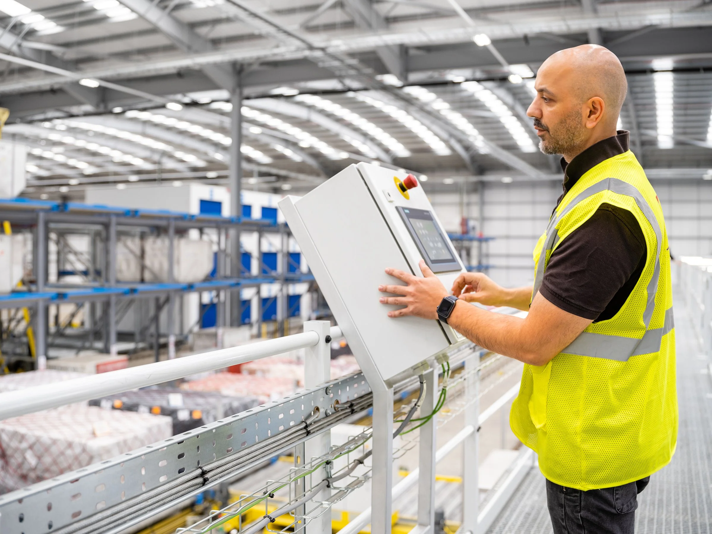 A man wearing a yellow safety vest and black shirt is standing in a large warehouse, operating a control panel with a touchscreen. The warehouse has metal shelving filled with boxes and goods, and overhead lighting illuminating the space.