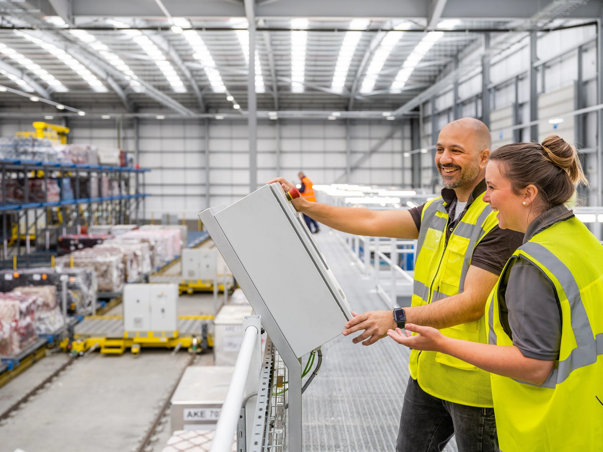 Two warehouse workers in yellow safety vests working on a control panel in a large industrial warehouse with shelves and pallets in the background.