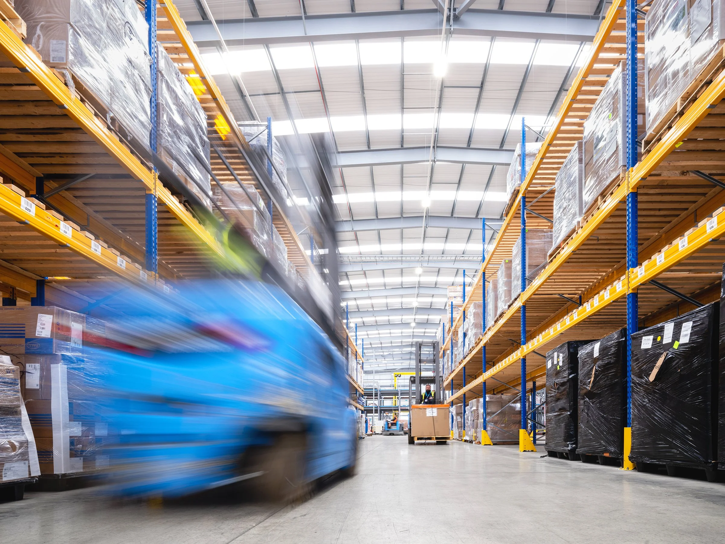 Interior of a warehouse with high shelves filled with palletized goods wrapped in plastic. A forklift is moving rapidly, causing motion blur, with a worker operating it in the background.