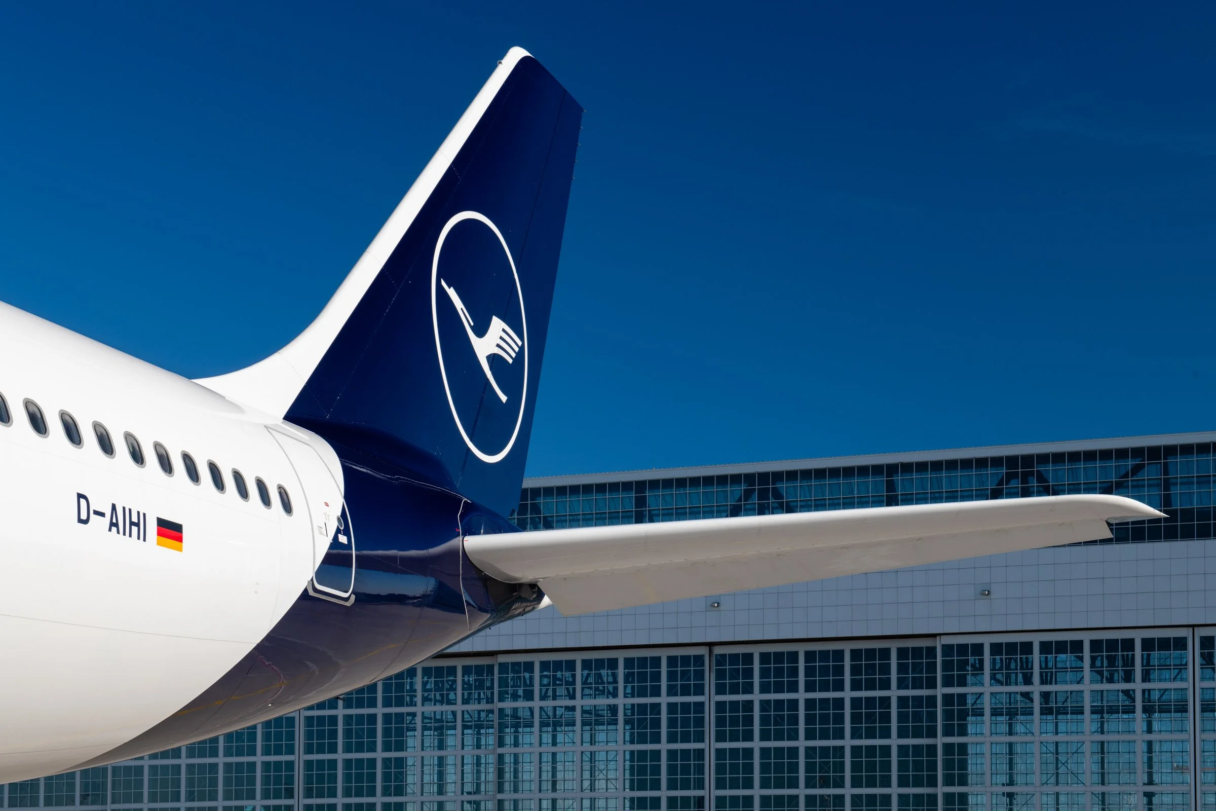 Close-up of a Lufthansa airplane tail with the airline's logo, against a modern glass building and blue sky.