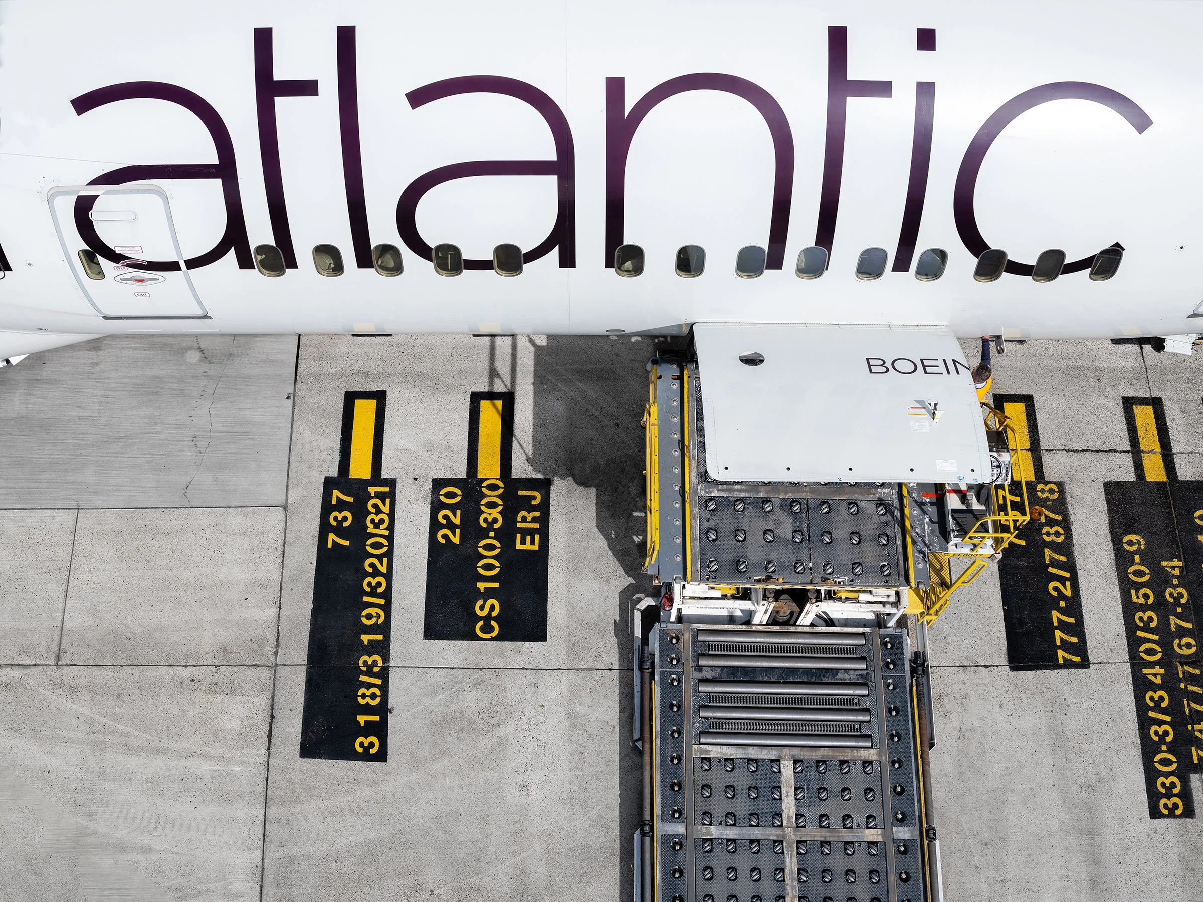 Overhead view of an airplane in the jet bridge, with an open door on the left side, connected to a jetway on the right, and yellow and black markings on the ground indicating parking positions.