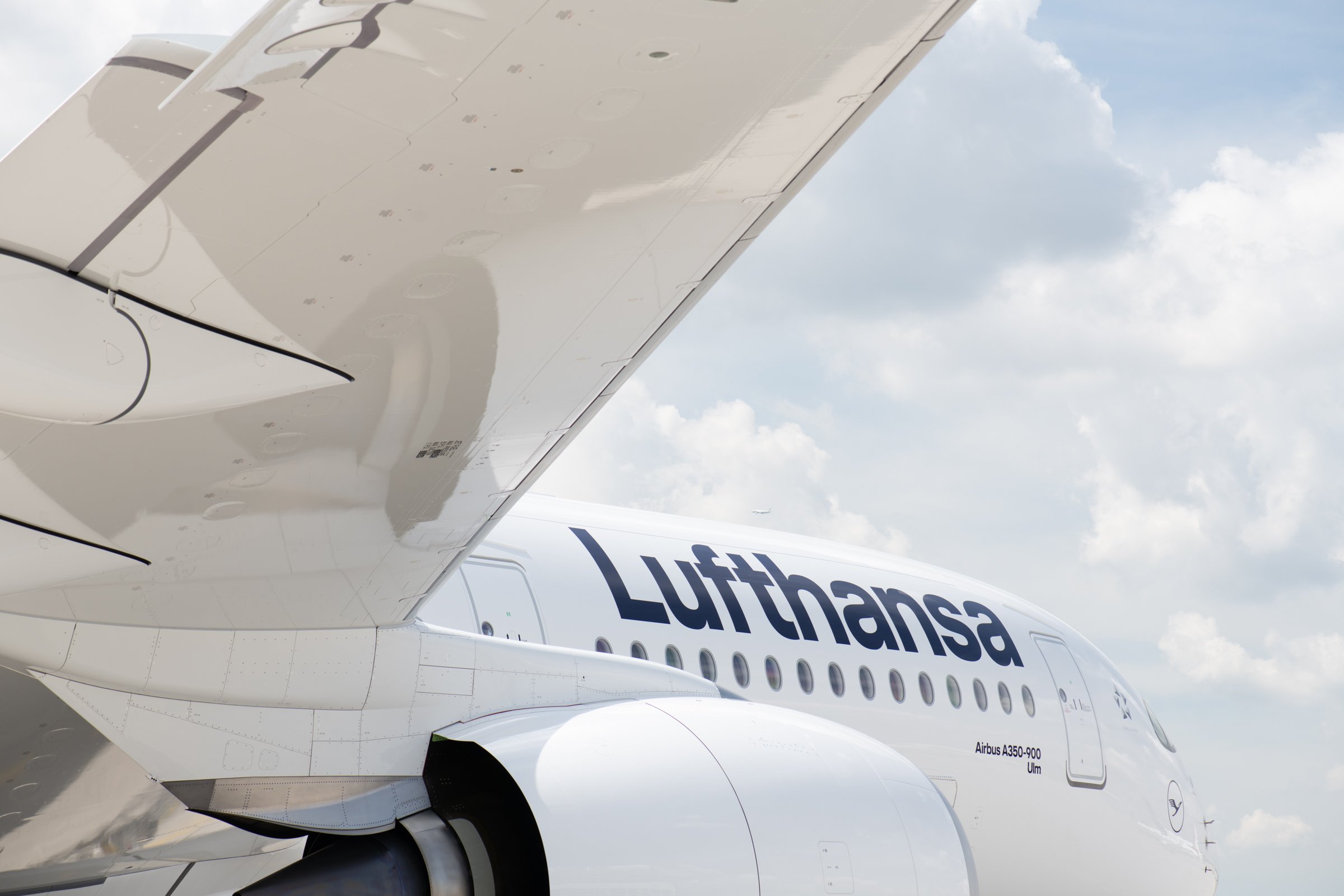 Close-up view of the underside of an Airbus A350 aircraft wing with Lufthansa branding, part of the fuselage and sky with clouds visible in the background.
