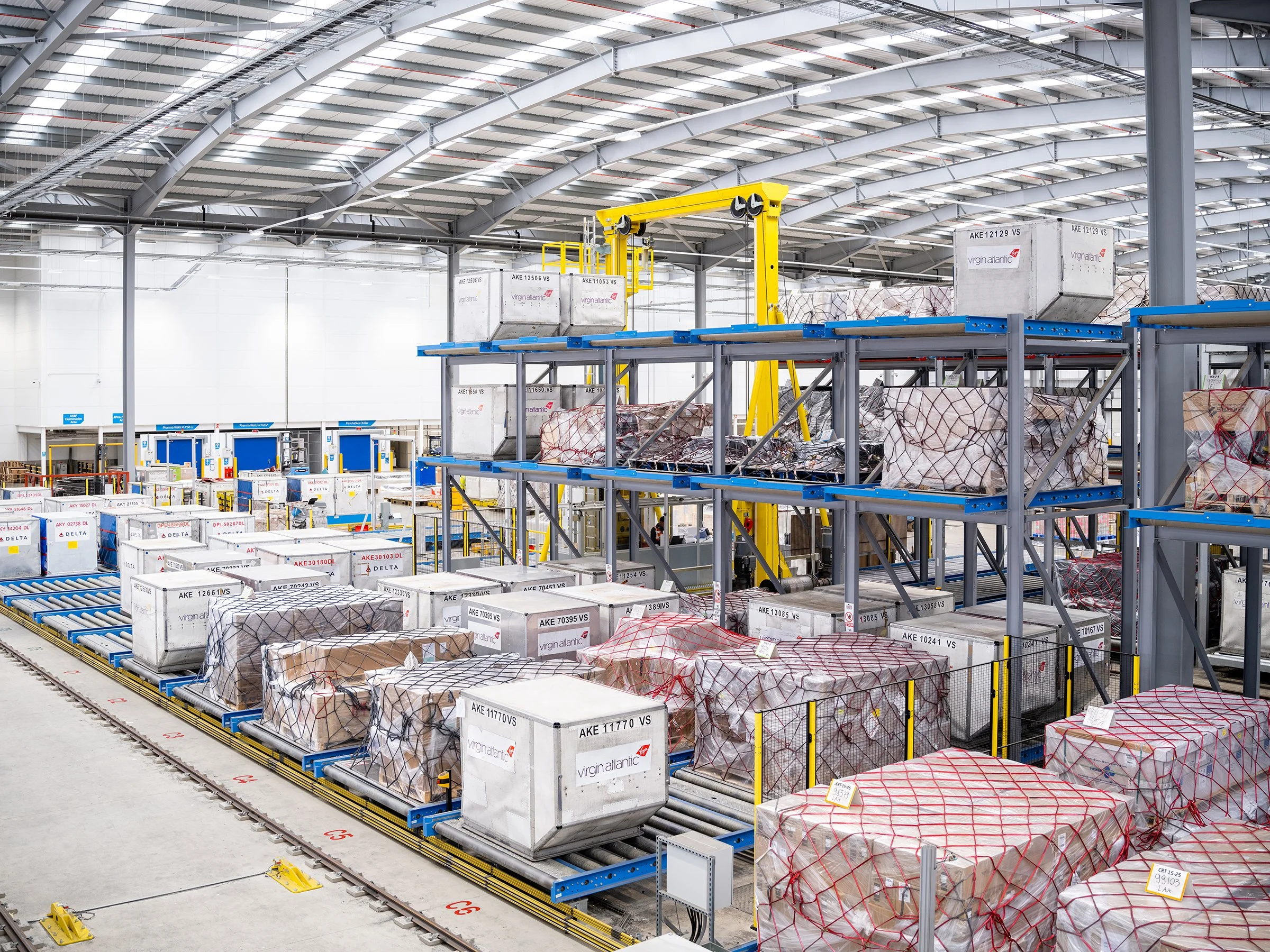 Inside a modern airport cargo terminal with organized shelves holding luggage and cargo containers, yellow and blue loading equipment, and a high arched metallic roof.