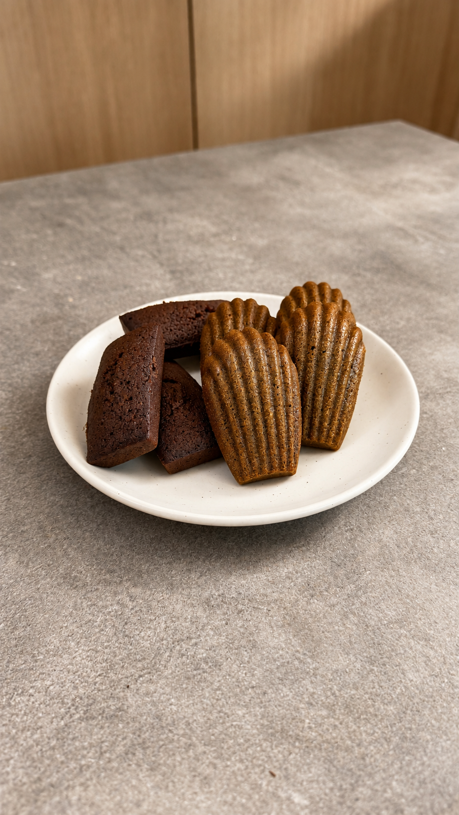 A white plate with two pieces of chocolate cake and three madeleines on a gray textured table.