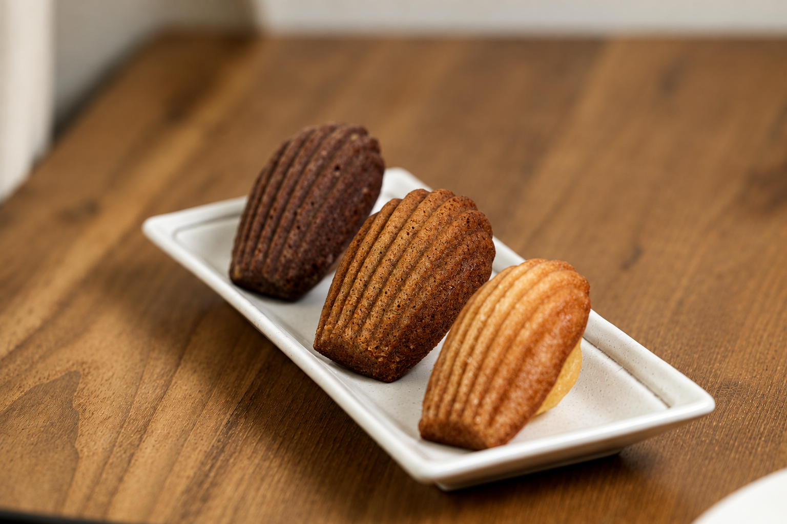 Three madeleine cookies, dark chocolate, cinnamon, and vanilla, on a white rectangular plate on a wooden surface.