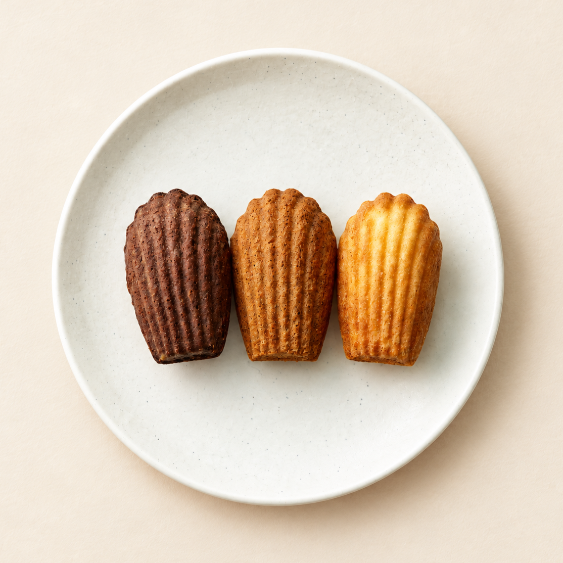 Three vanilla and chocolate mini madeleines on a white round plate against a light beige background.