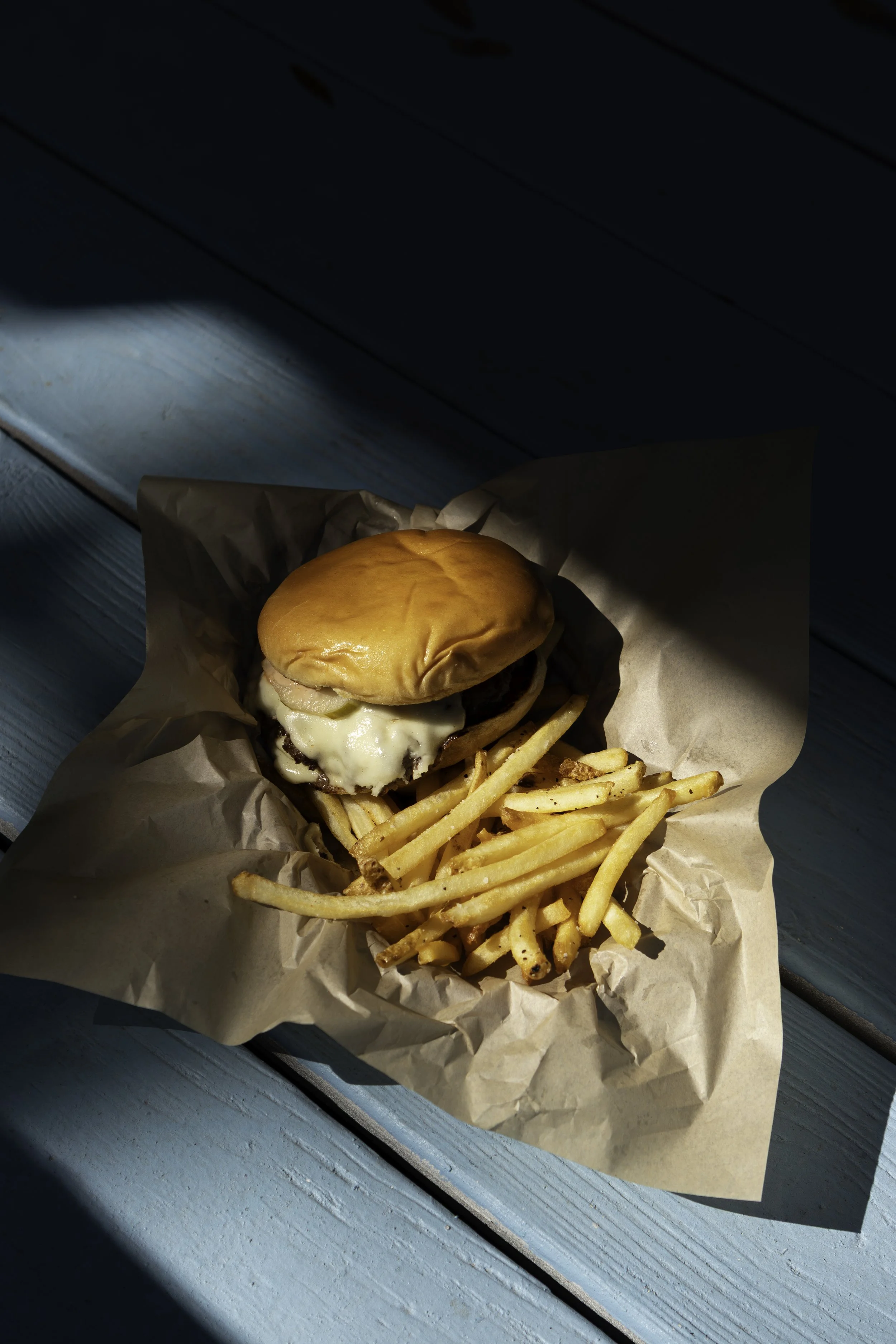 Cheeseburger with fries on crumpled paper, on a blue wooden surface with sunlight shadows.