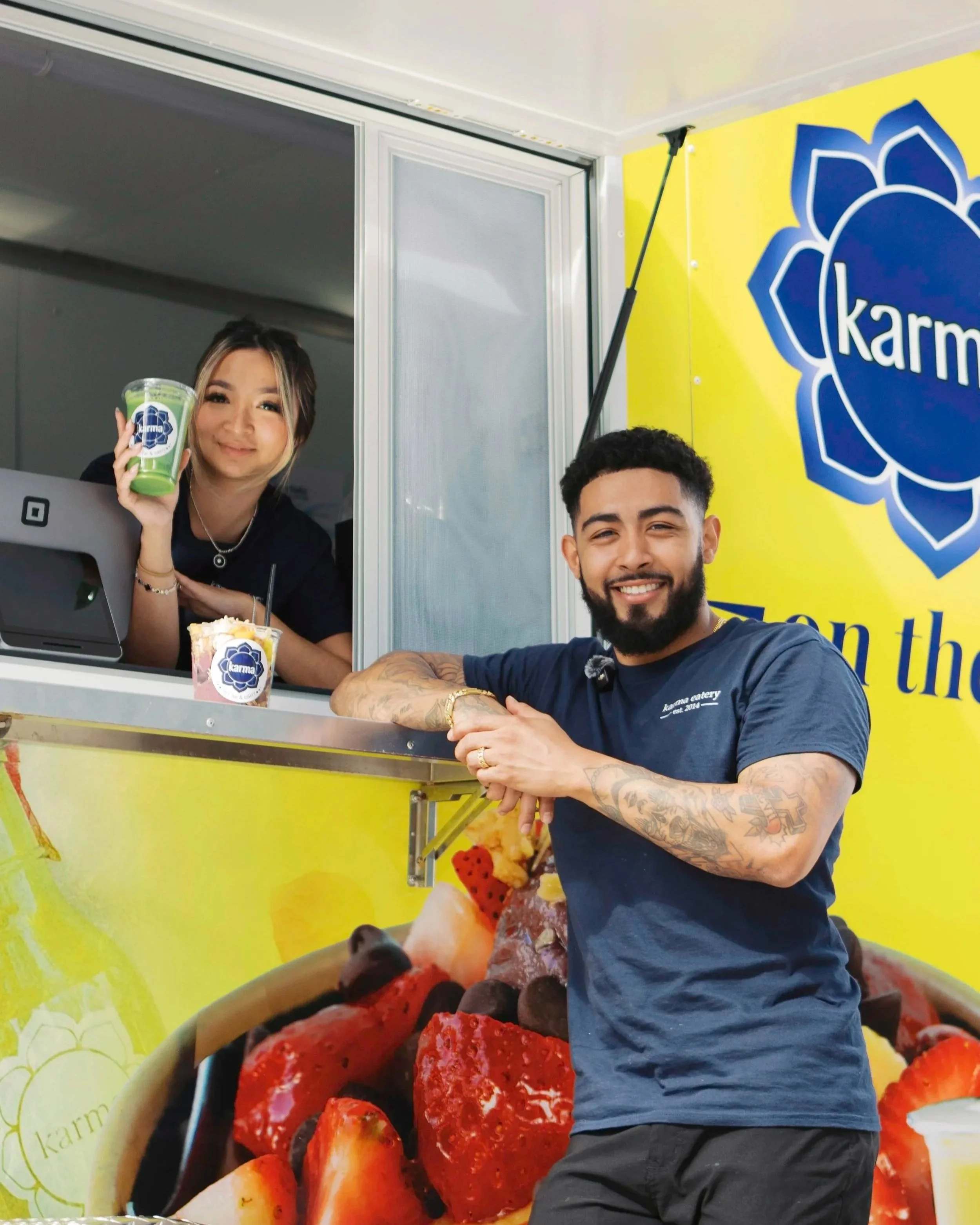 A woman and a man are at a colorful food truck with a yellow background and strawberries. The woman is inside the food truck, holding a green drink or dessert cup with the Karma logo, smiling at the camera. The man is standing outside, smiling at the