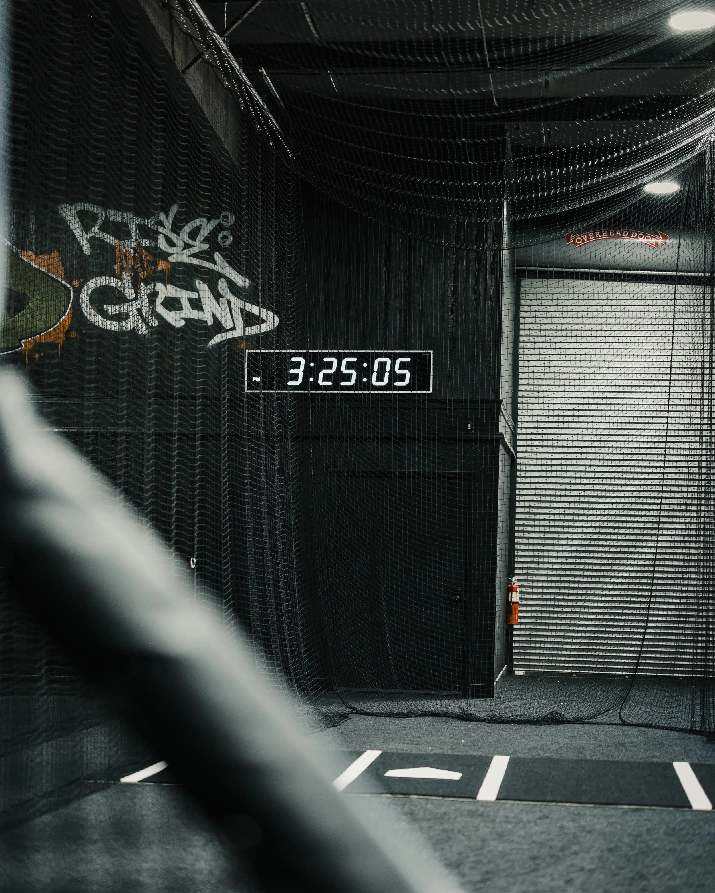 Indoor batting cage with black netting and a digital clock showing 3:25:05. Sign on the back wall reading 'Overhead Door'.