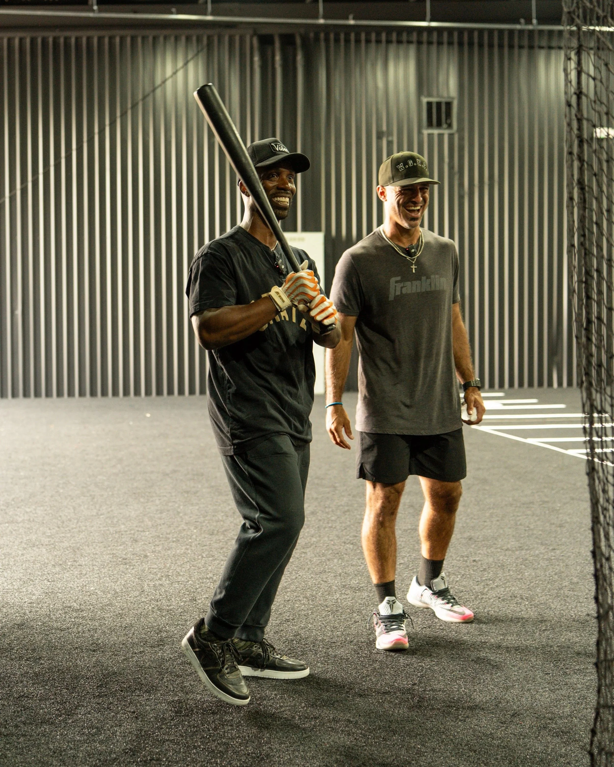 Two men walking together in an indoor facility, both dressed in athletic wear, smiling and laughing.