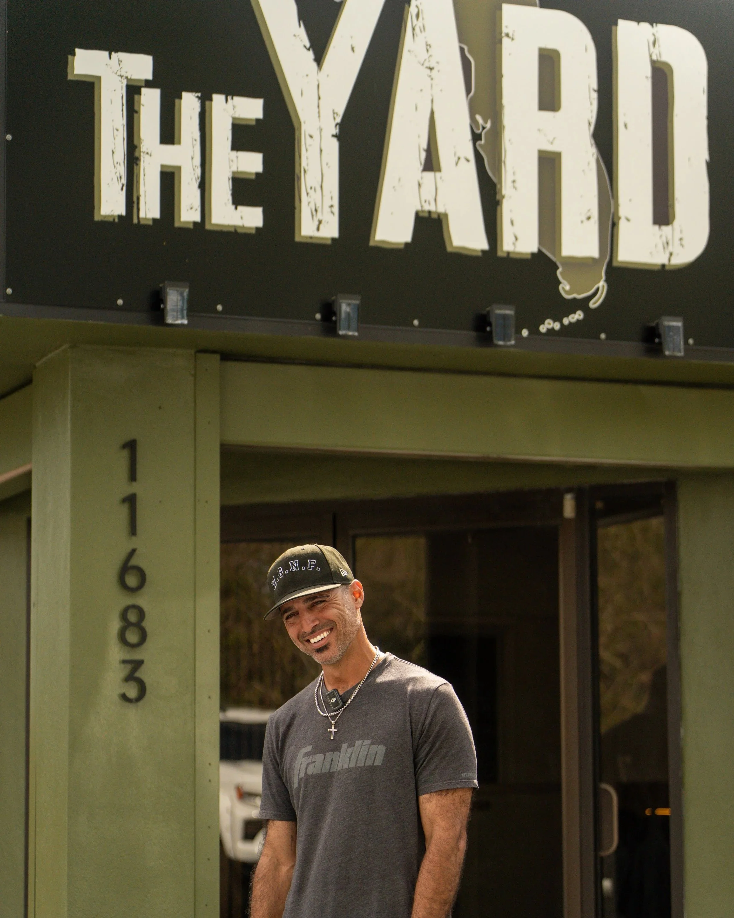 A smiling man wearing a baseball cap and a cross necklace standing outside near a green clock structure with large white Roman numerals. Part of the words 'THE YARD' can be seen above him.