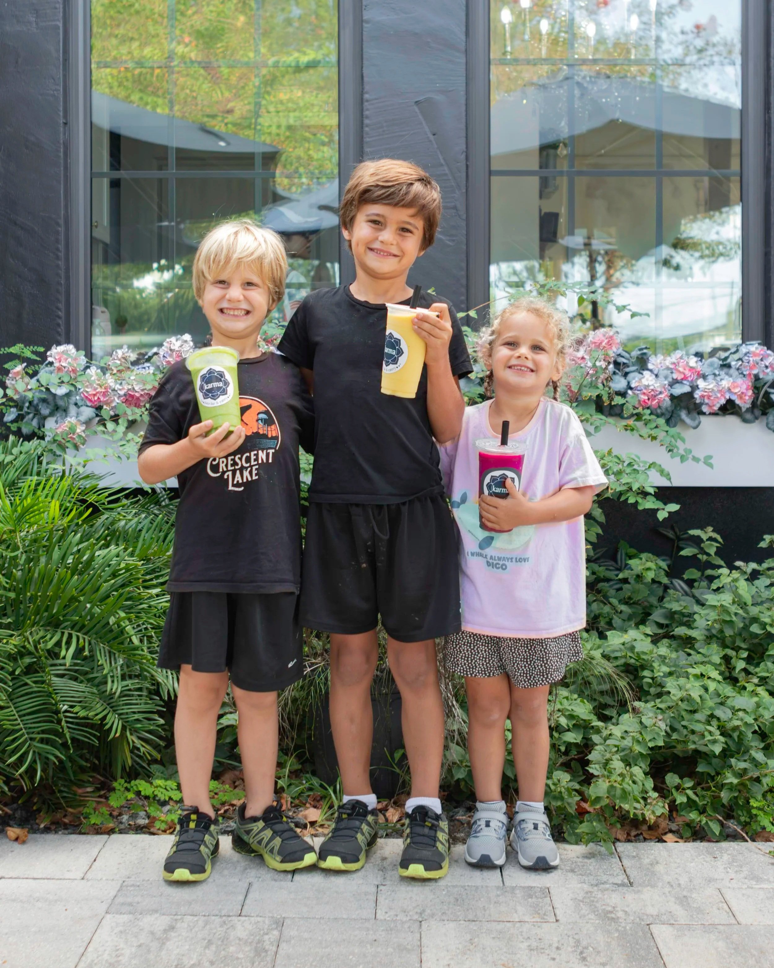 Three smiling children standing outdoors in front of a windowed building, holding smoothies, surrounded by greenery.