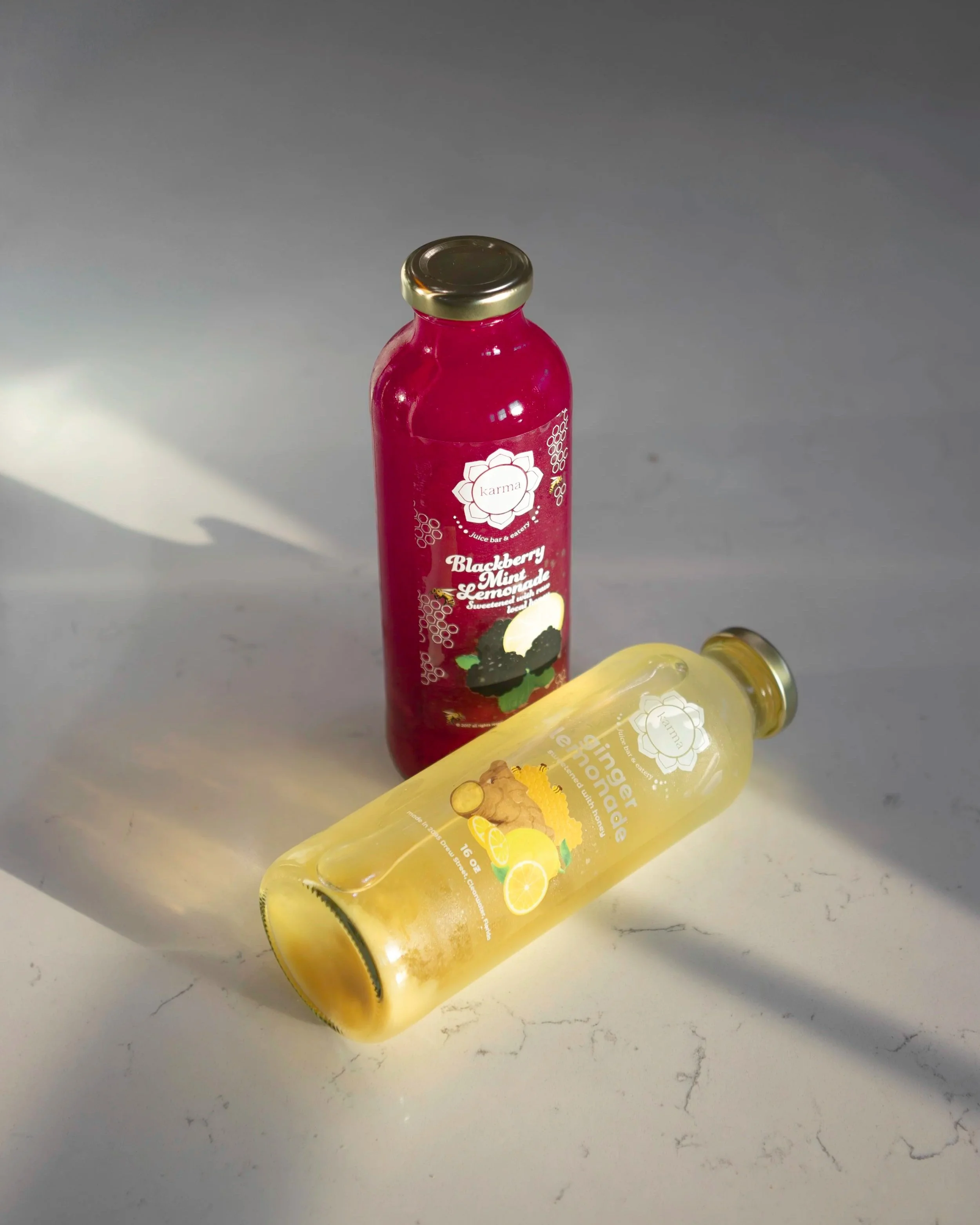 A red bottle of Karma blackberry mint lemonade and a yellow bottle of Karma ginger lemonade resting on a white marble surface.