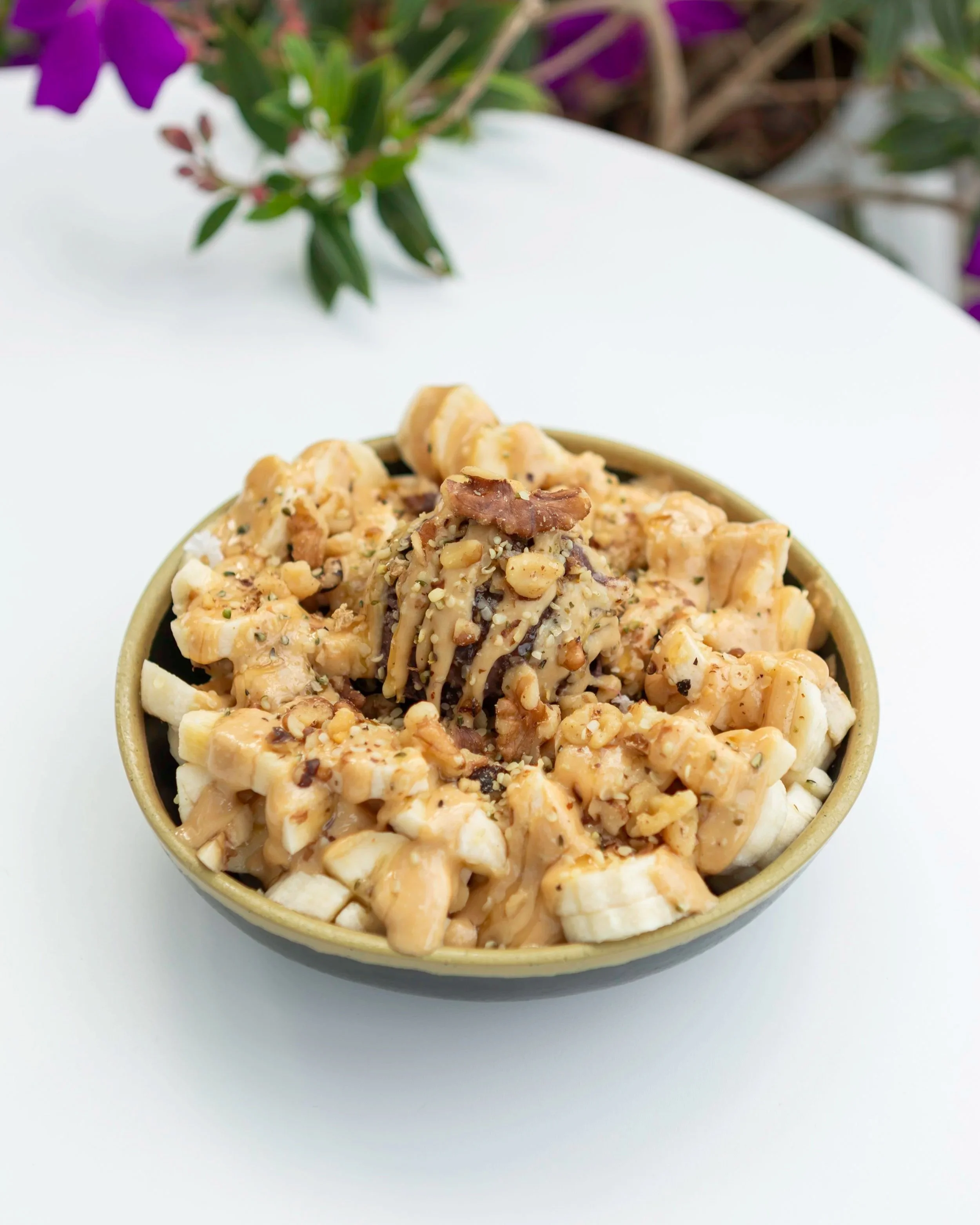 A bowl of macaroni and cheese topped with chopped nuts, served on a white surface with purple flowers in the background.