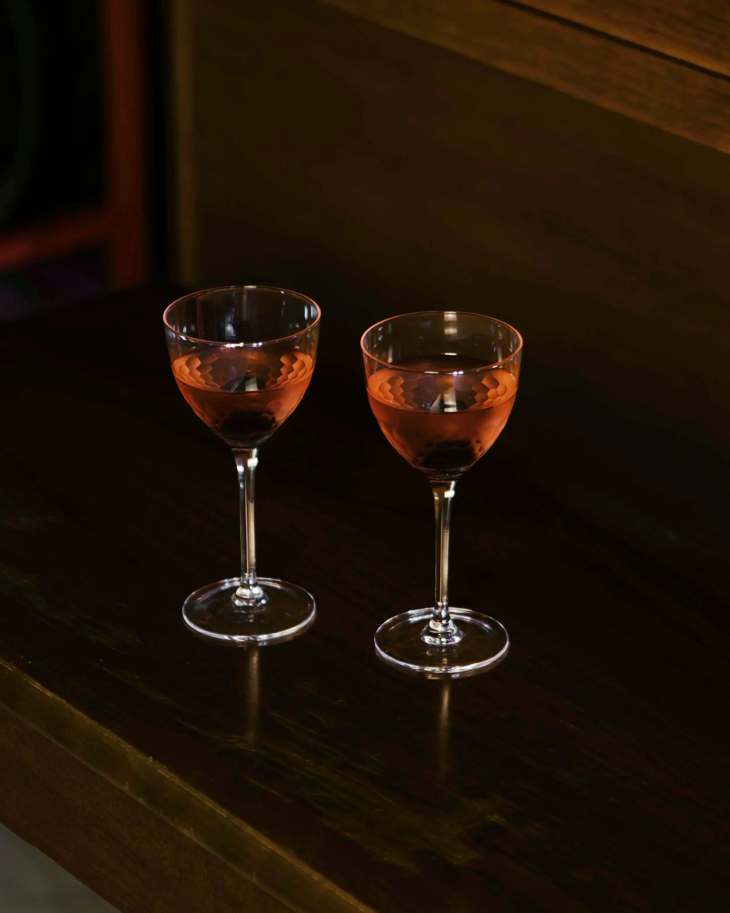Two pink-colored glasses filled with a clear liquid, placed on a dark wooden table in a dimly lit setting.