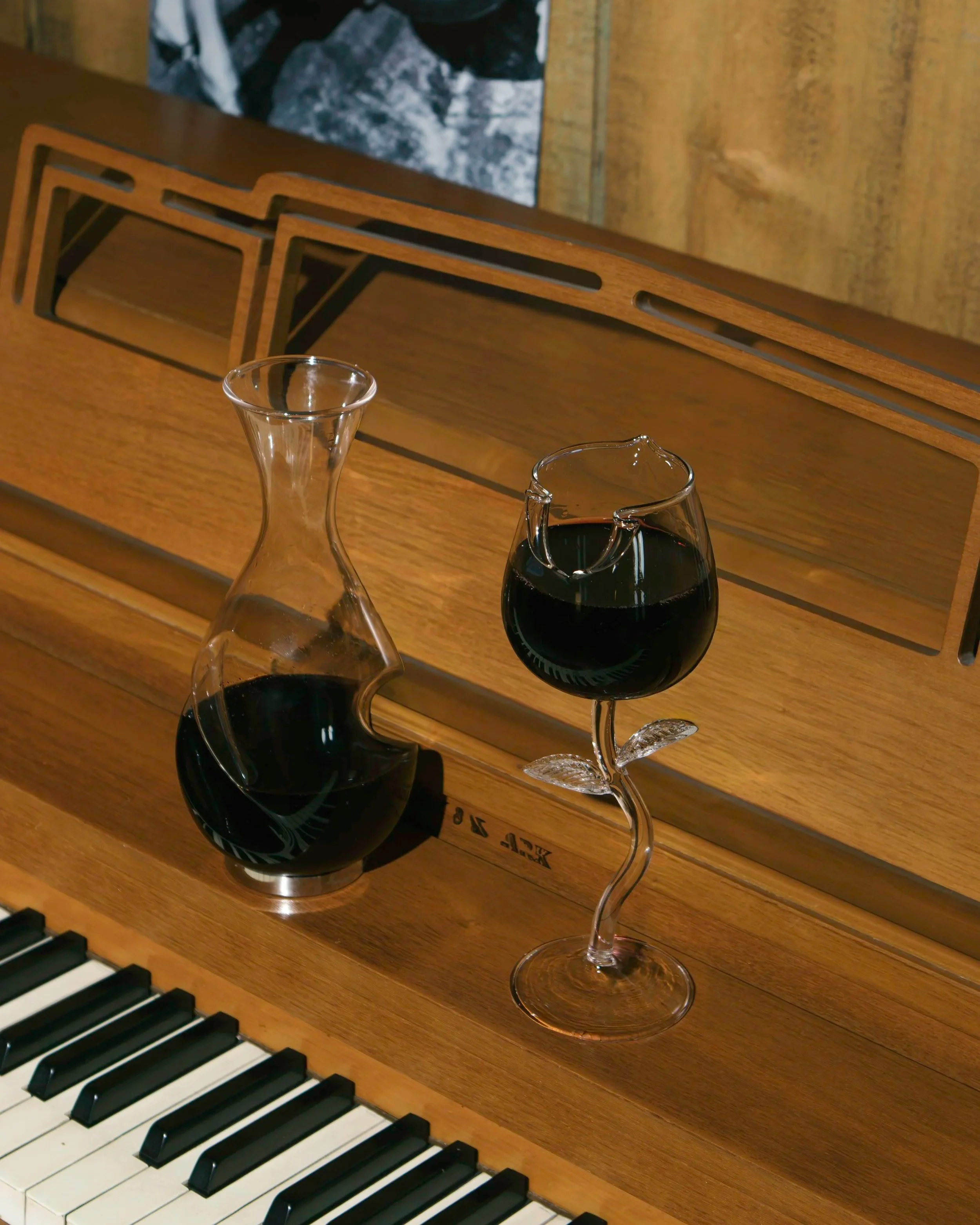 A wooden piano with white and black keys, a glass decanter, and a wine glass filled with red wine, with two matching glasses behind. The background shows a wooden panel and a piece of artwork.