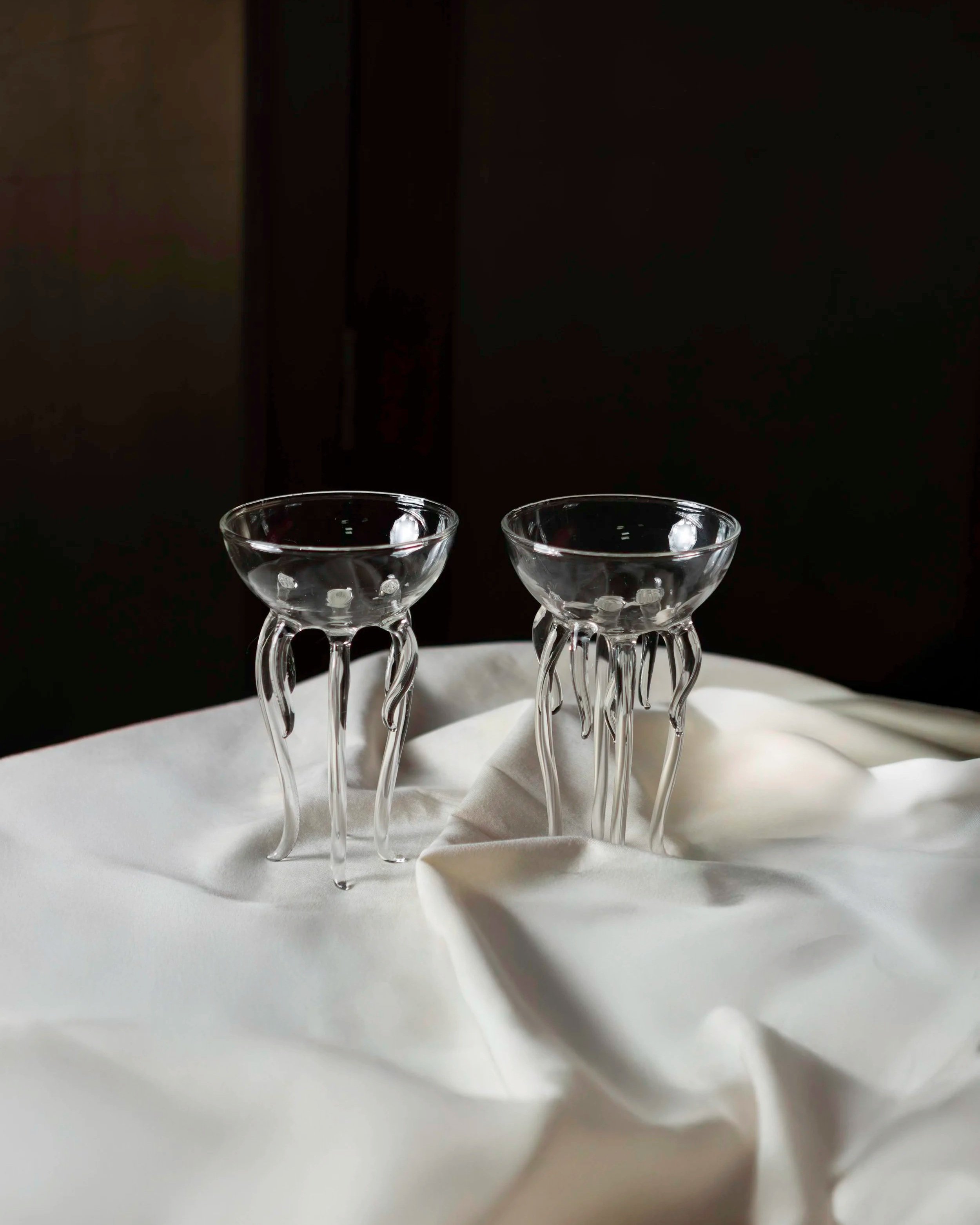 Two clear glass goblets with swirled stems placed on a white fabric surface against a dark background.